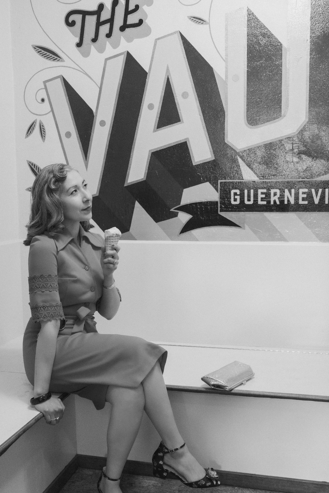 A woman sits on a bench holding an ice cream cone, gazing thoughtfully at a mural that reads "THE VAULT" on the wall behind her.