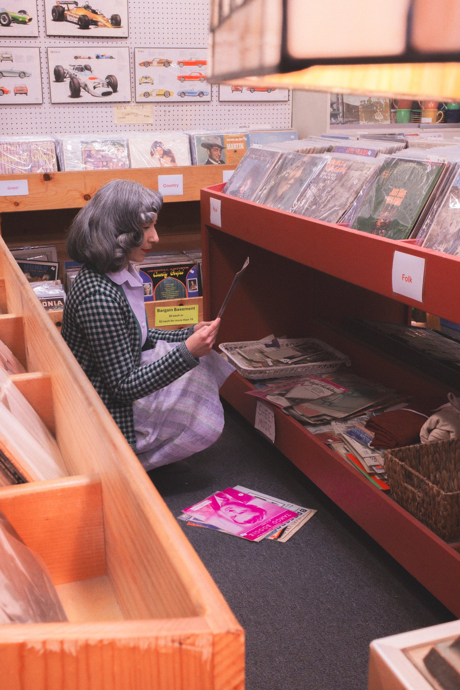 Sheet Music Stories gallery image: A woman in a checked blazer examines sheet music while crouching beside a low shelf filled with albums in a vintage-style record store.