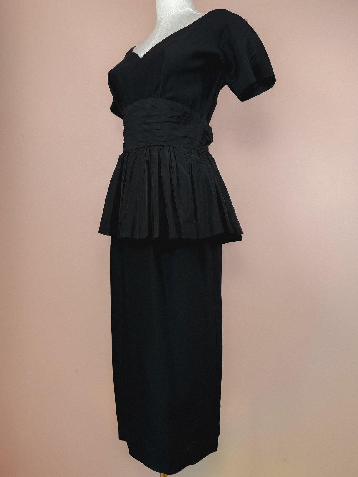 Black 1950s women’s dress featuring a fitted bodice and pleated peplum detail.