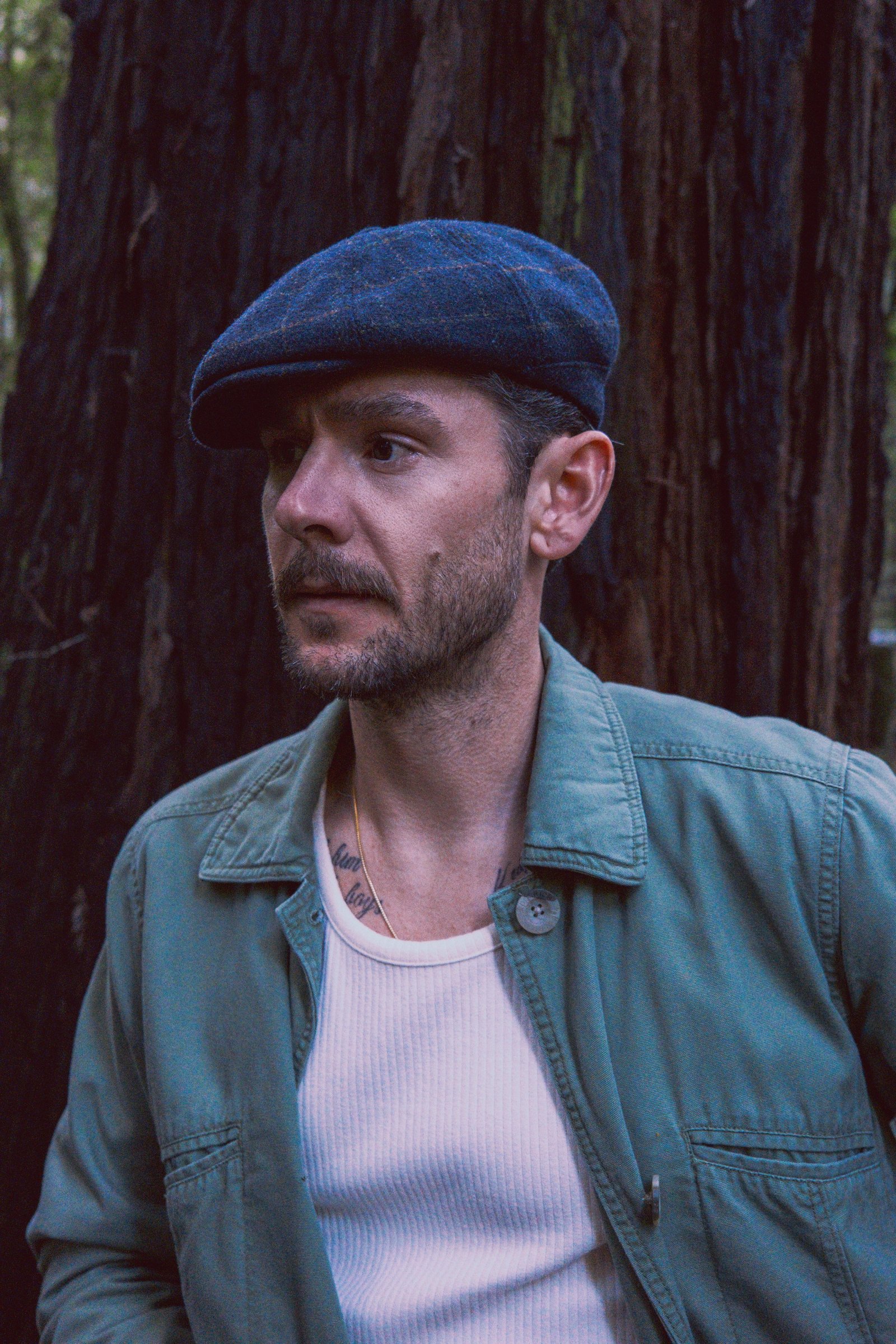 A man with short hair wearing a flat cap and a green jacket stands near a tree in a forest.