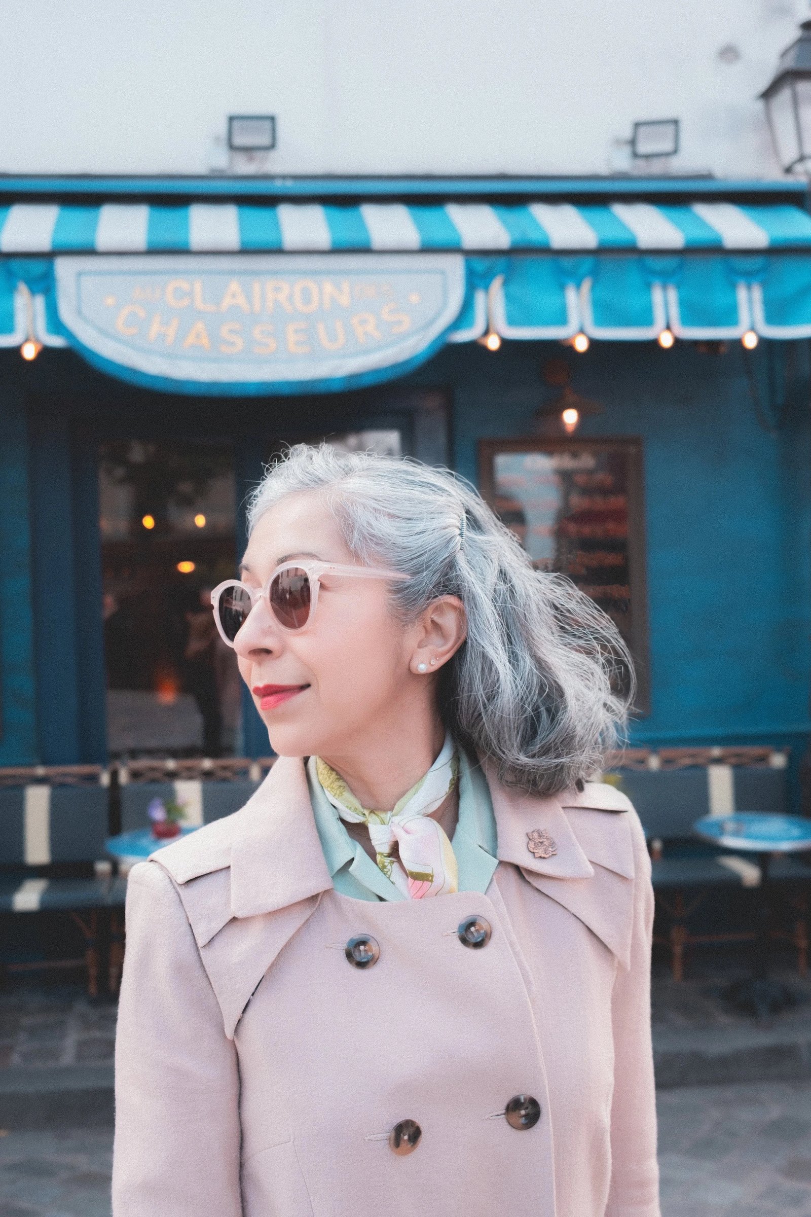 A woman with silver hair wearing sunglasses and a pastel coat stands outside a café with blue and white striped awnings.