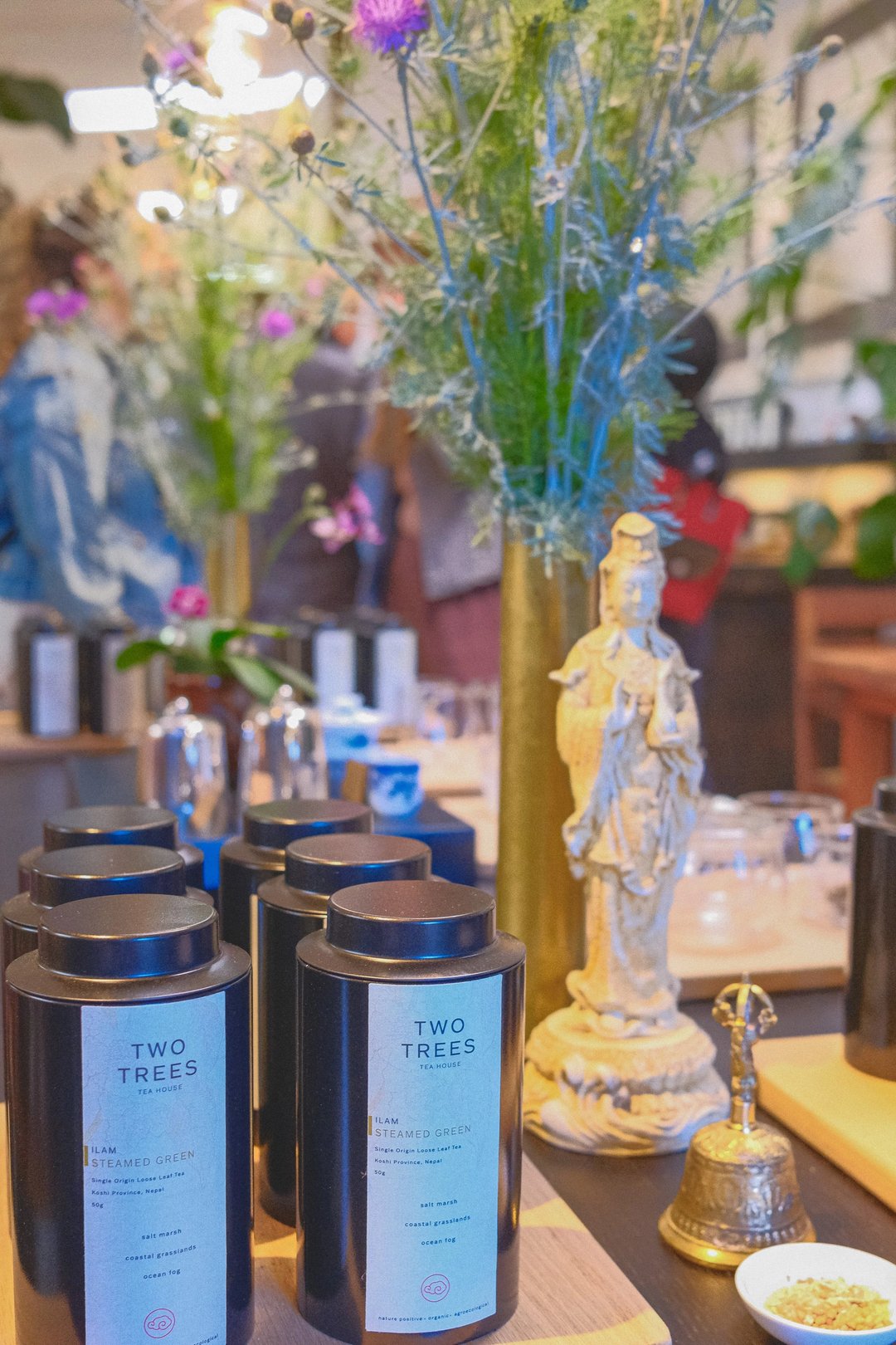 A display of black candle jars from "Two Trees" alongside a decorative statue and vibrant floral arrangements in a retail space.