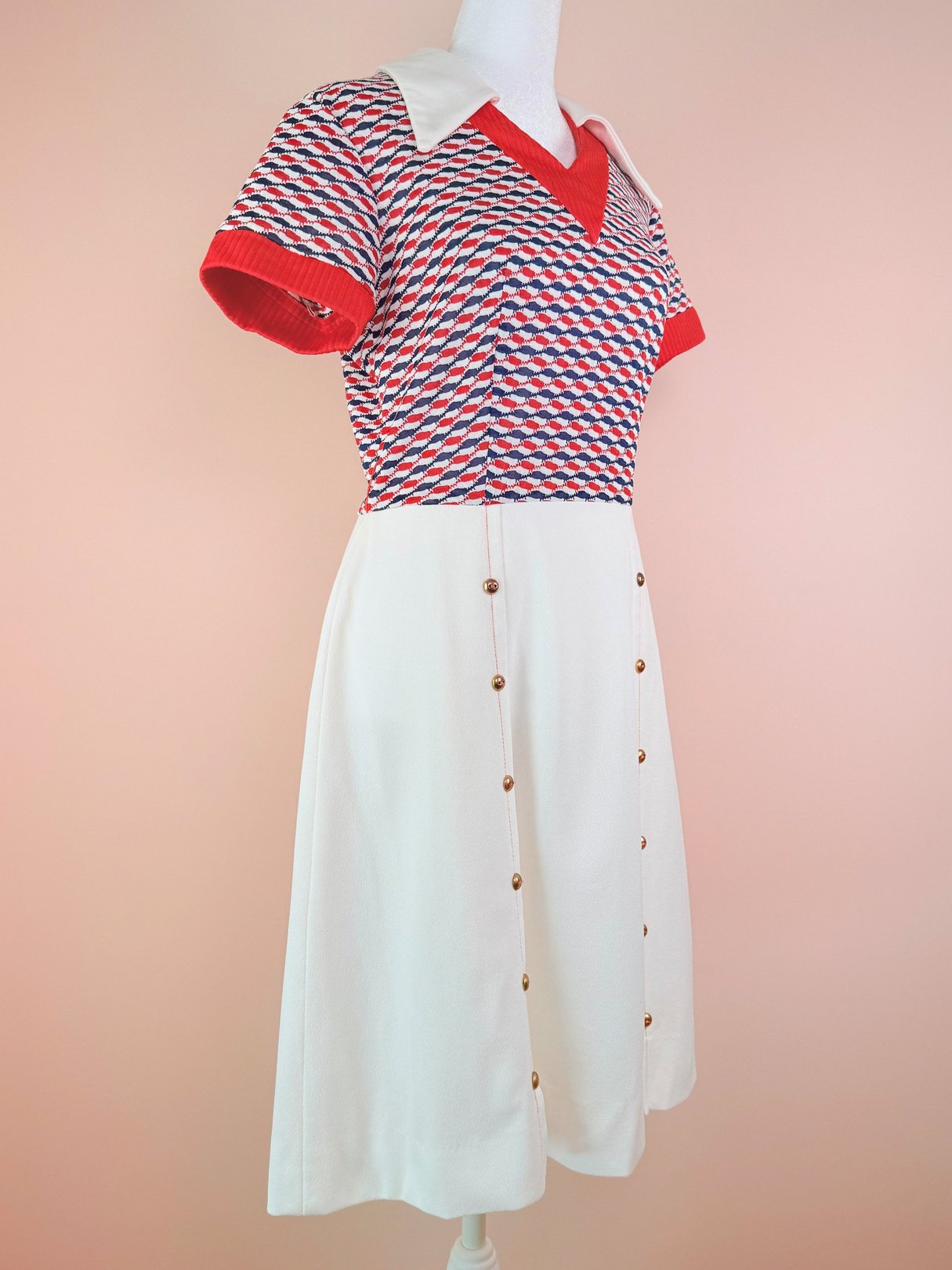 The Bottle Rocket Dress features a red and patterned top with short sleeves and a white skirt adorned with buttons, representing 1970s style for women.