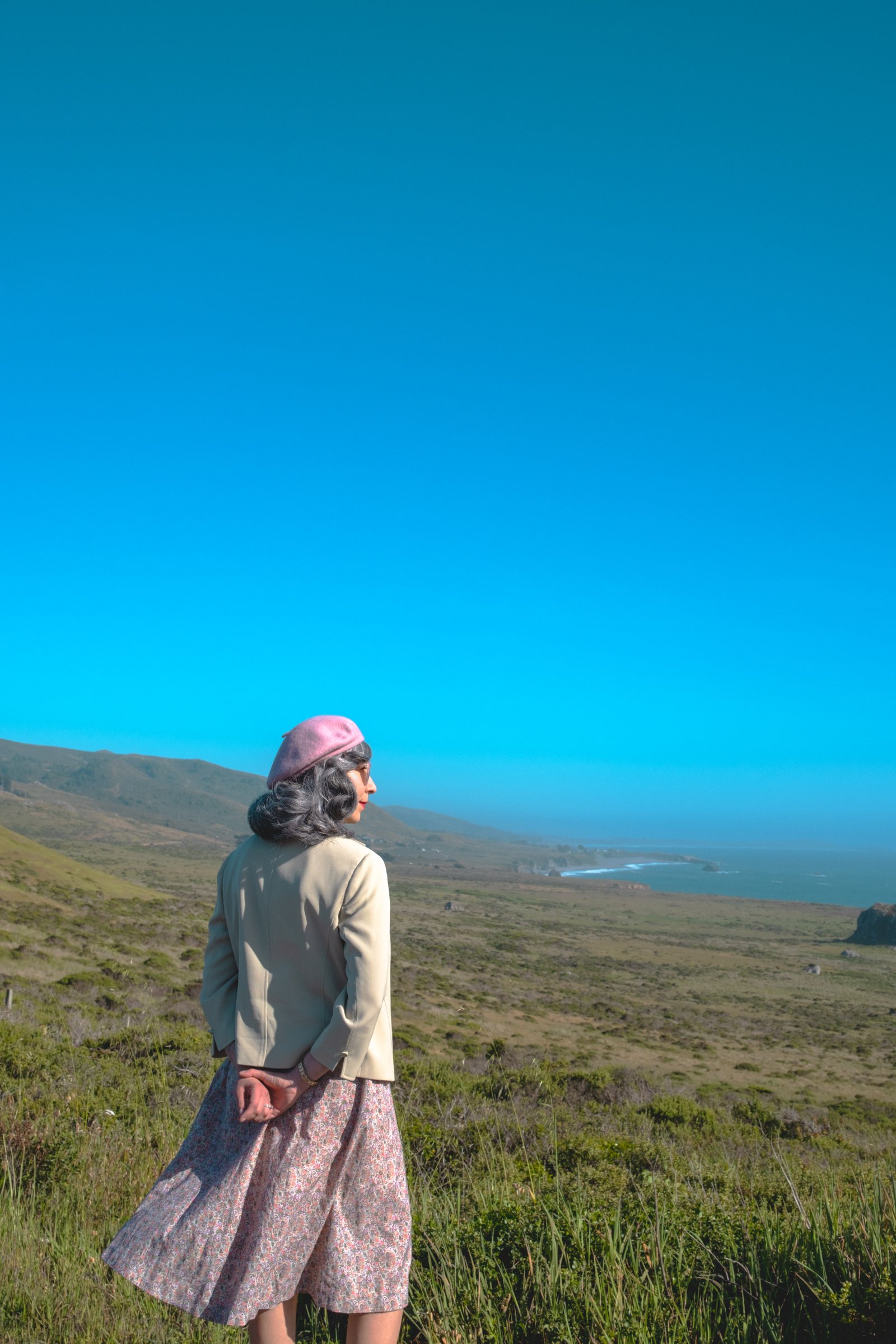 The Road to Bodega Bay gallery image: A woman in a vintage outfit stands overlooking a coastal landscape with hills and the ocean in the distance under a clear blue sky.