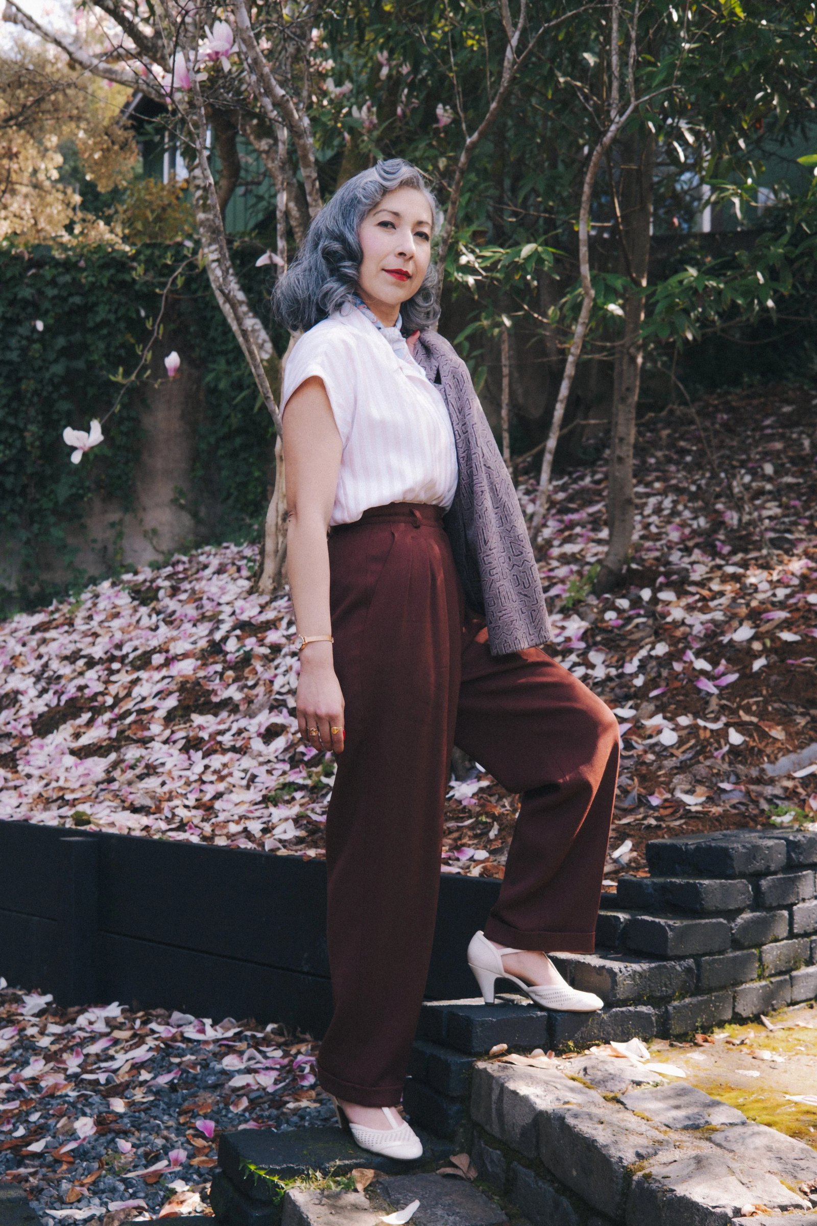 A woman with gray hair stands on stone steps in a garden, wearing a white blouse, burgundy pants, and white heels, surrounded by fallen petals.