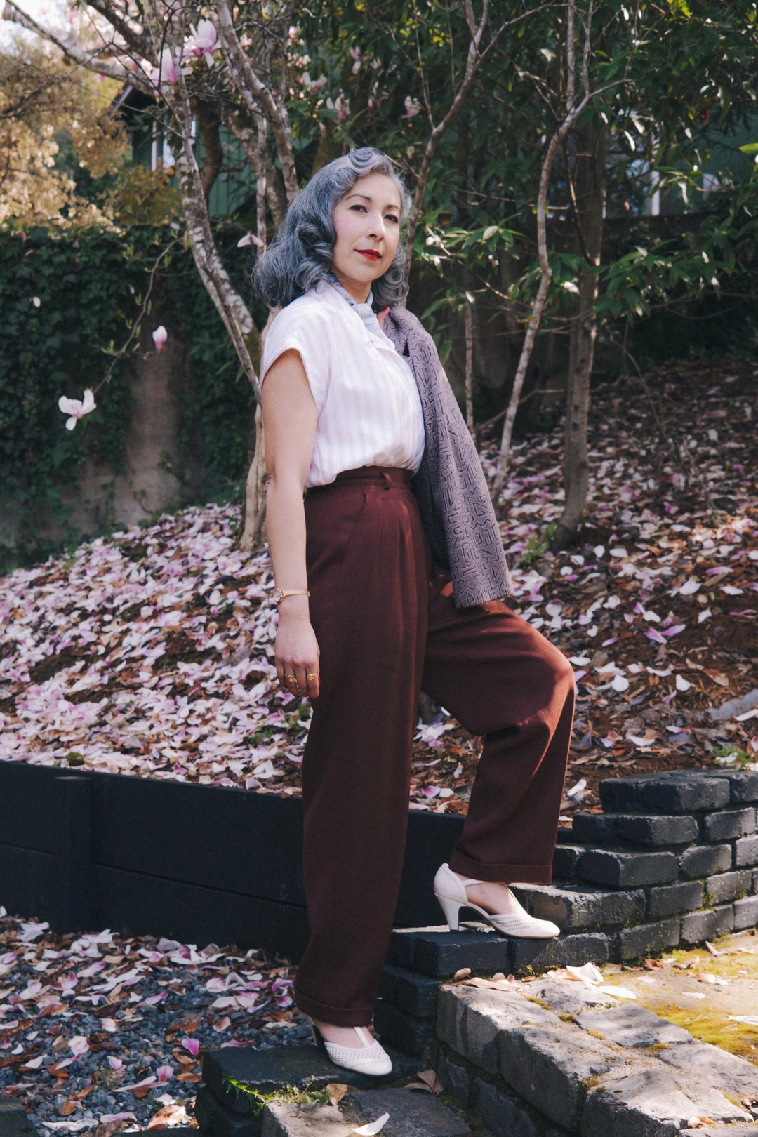 A woman with gray hair stands on stone steps in a garden, wearing a white blouse, burgundy pants, and white heels, surrounded by fallen petals.