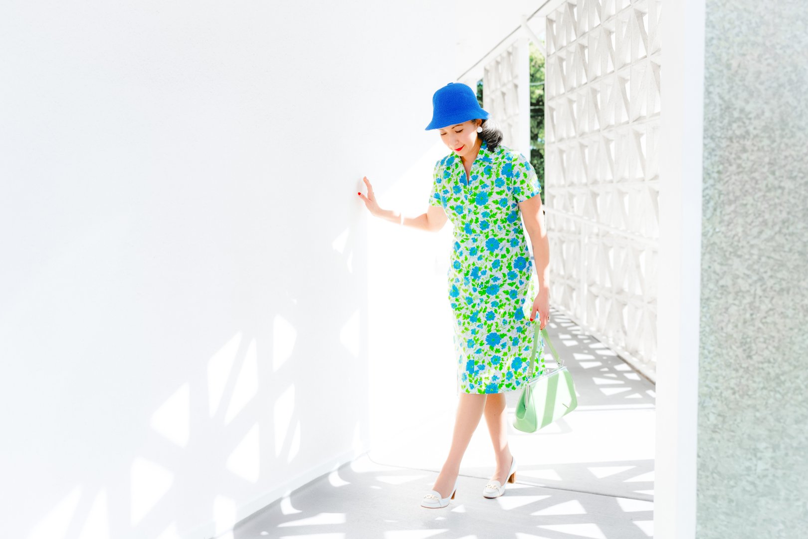 A woman dressed in a floral green and blue dress, paired with a blue hat and carrying a green bag, walks through a bright, modern corridor.
