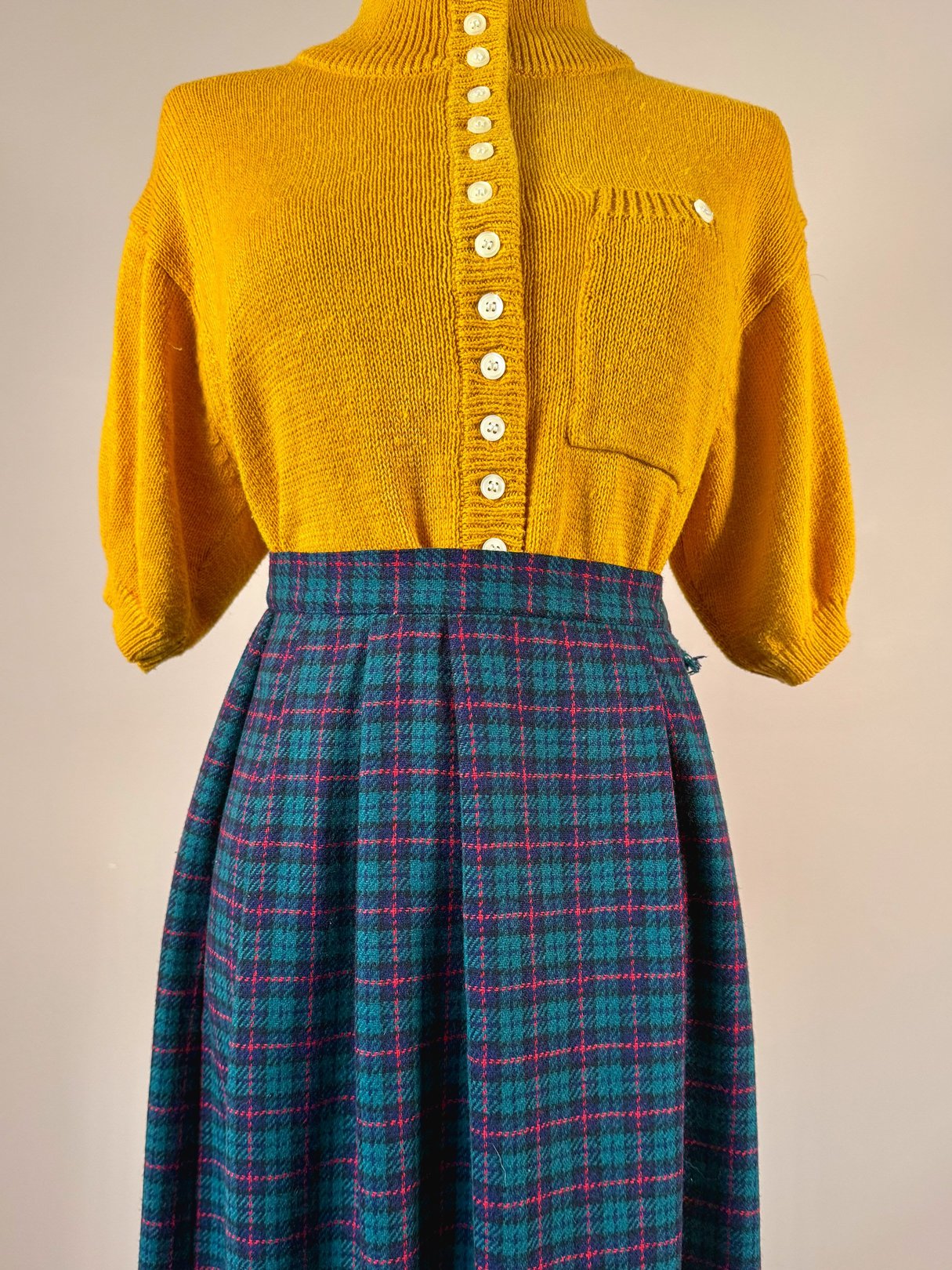 A 1970s women's green plaid skirt featuring a fitted waist and a colorful check pattern, paired with a mustard yellow knit sweater.