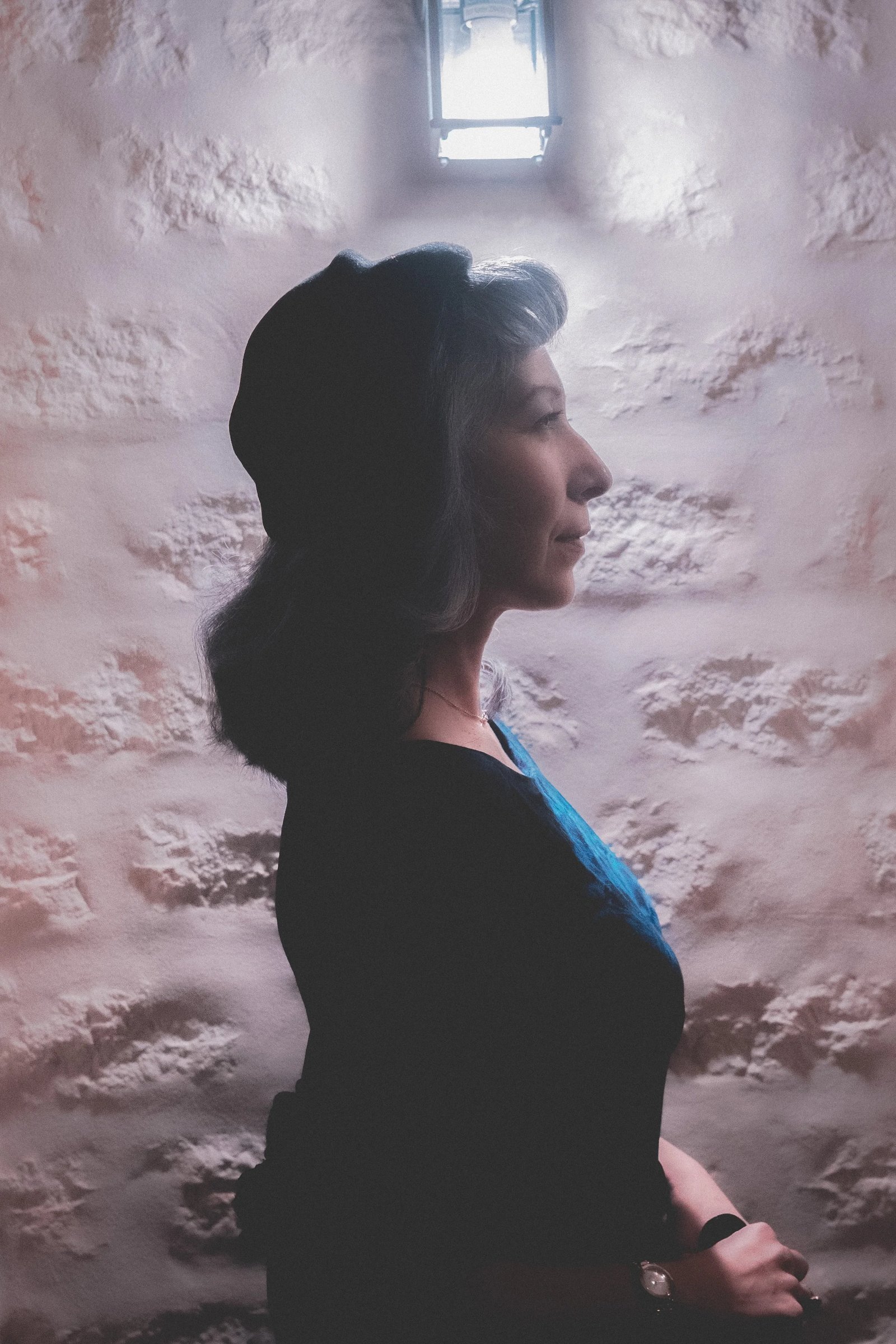 A woman wearing a black hat and a blue top is shown in profile against a textured wall.
