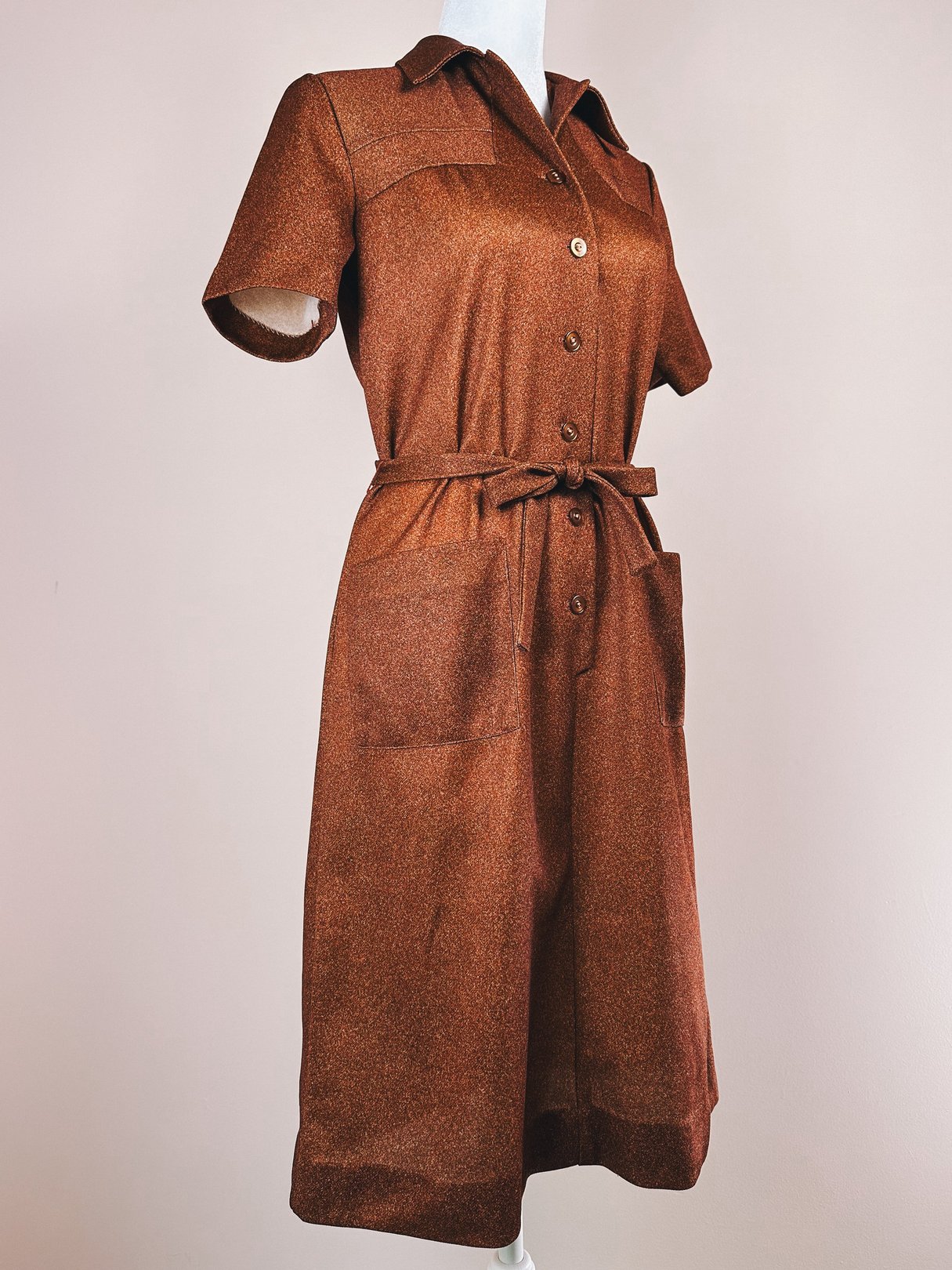 A 1970s women's brown day dress featuring short sleeves, a collar, front button closure, and a tie belt with two front pockets.