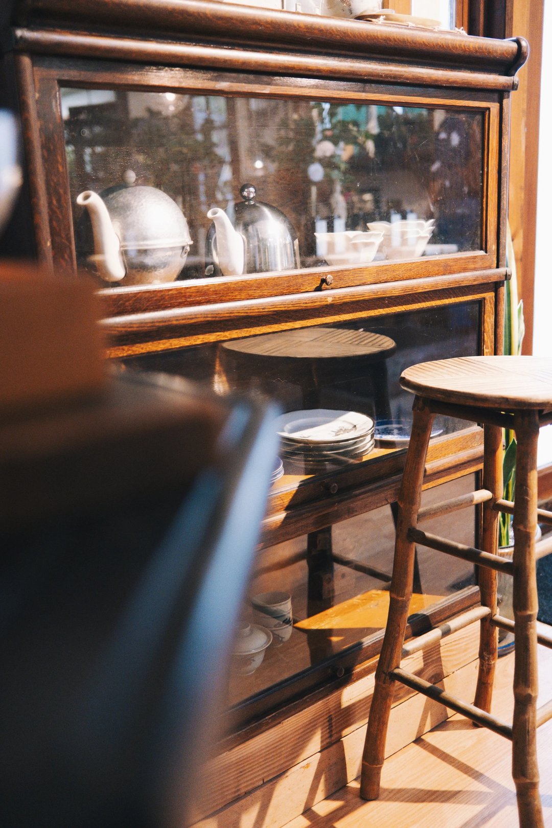 A vintage wooden display cabinet showcasing tea sets and ceramic plates, with a wooden stool in the foreground.
