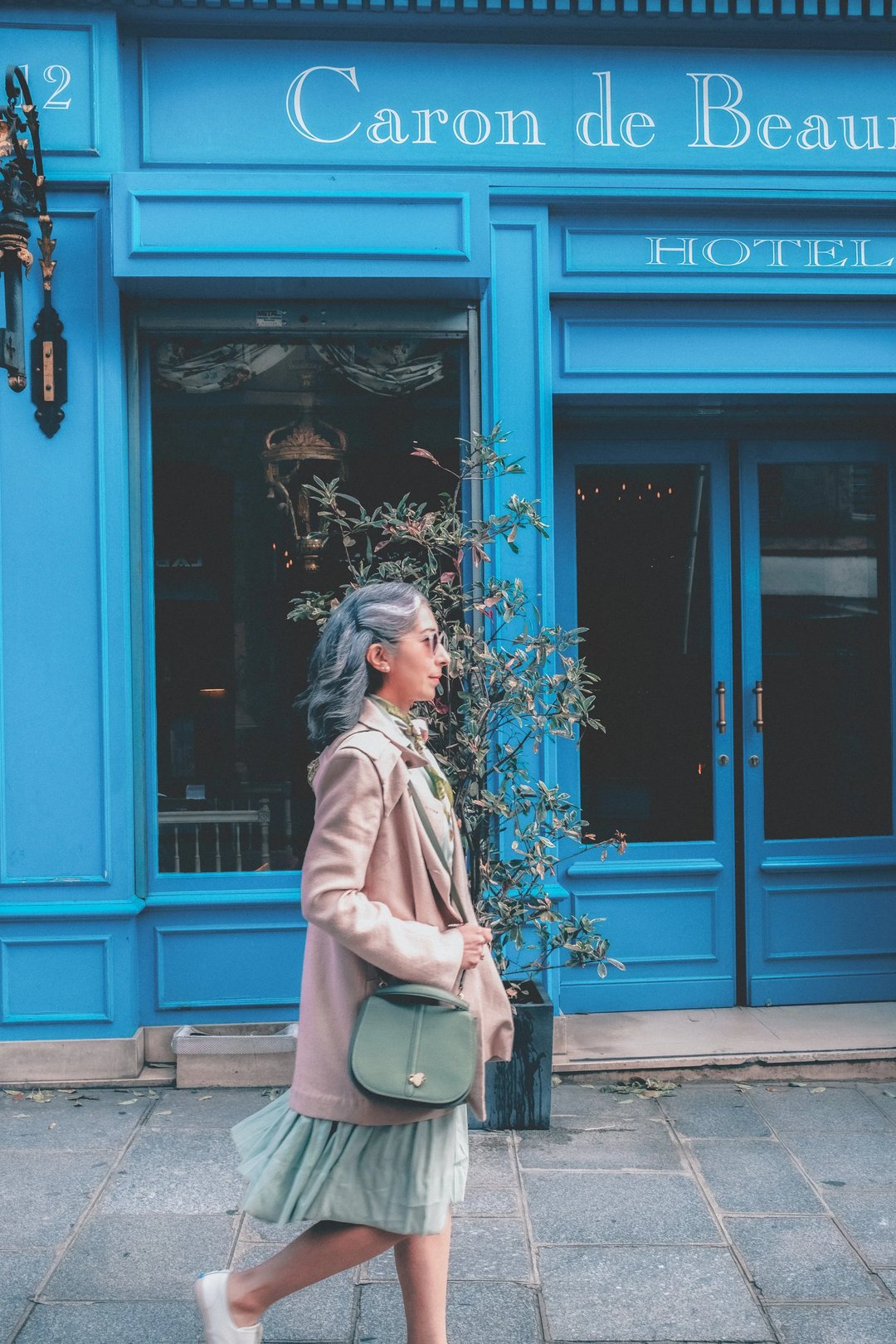 A woman with gray hair walks past a bright blue building, dressed in a beige coat and green skirt, carrying a small handbag.