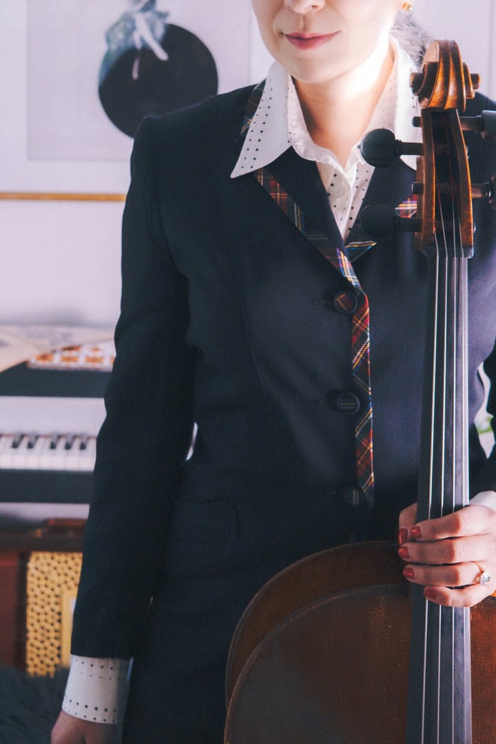 A person in a tailored black suit with a plaid tie stands beside a cello, set against a backdrop of musical instruments.