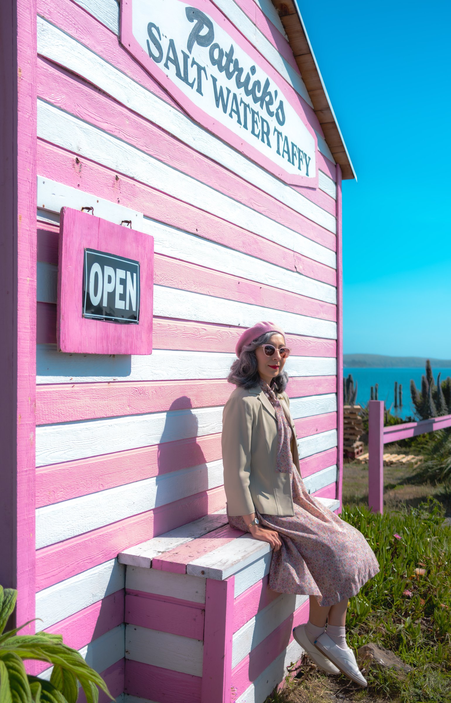 The Road to Bodega Bay gallery image: A woman in a vintage dress and pink accessories sits on the steps of a pastel pink building labeled "Patrick's Salt Water Taffy," with a coastal view in the background.