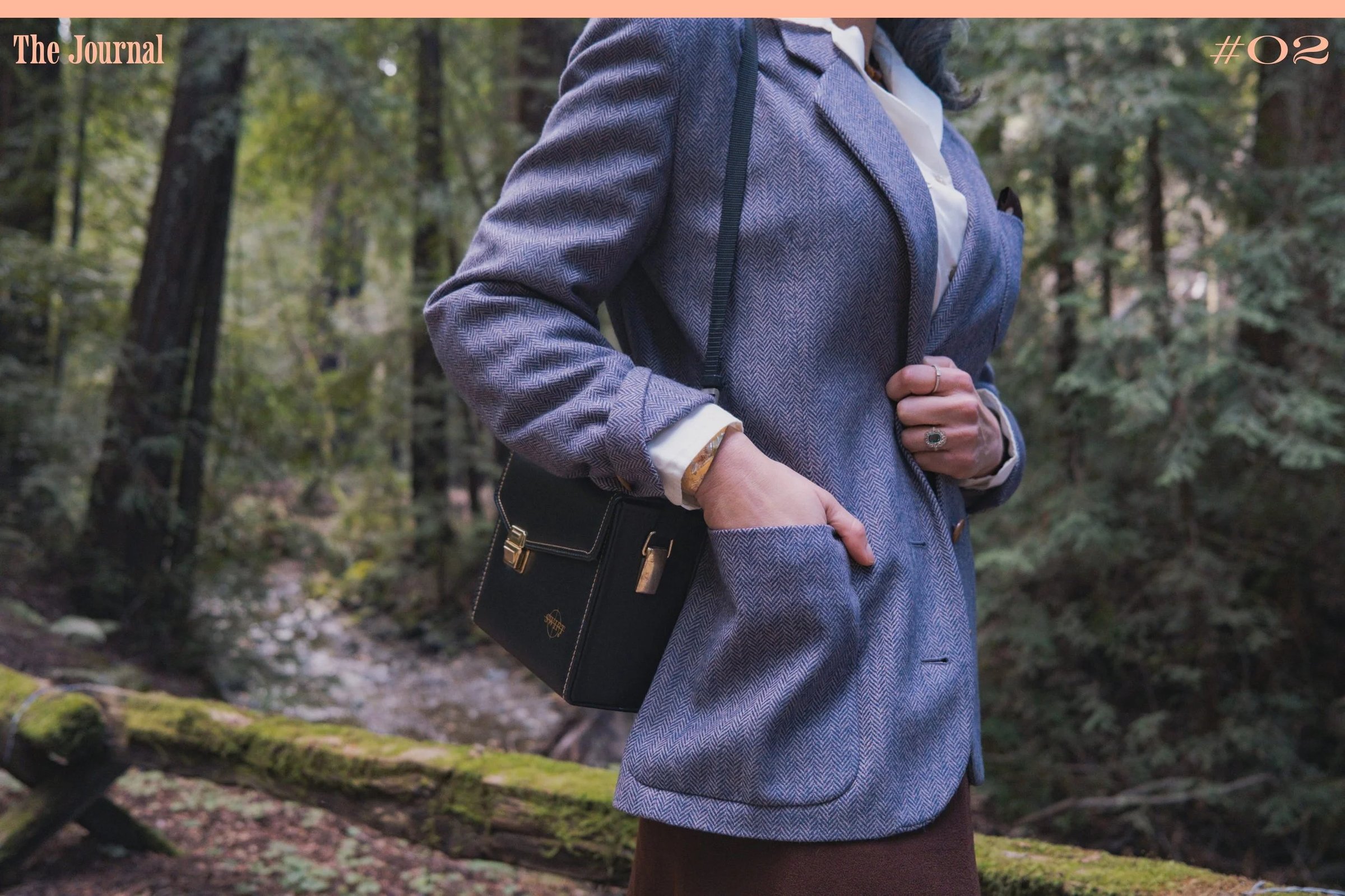 A person in a herringbone blazer holds a small black handbag in a natural outdoor setting.