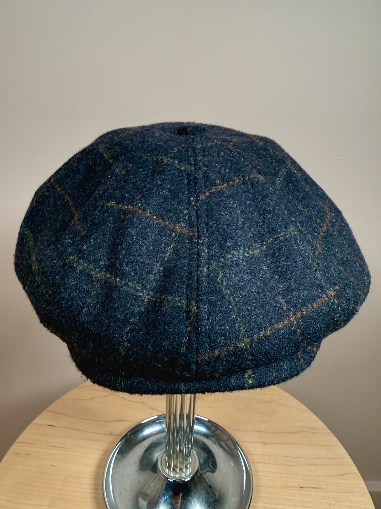 Navy blue tweed cap with a vintage pattern, designed for men, reflecting 1970s style, displayed on a metal stand.