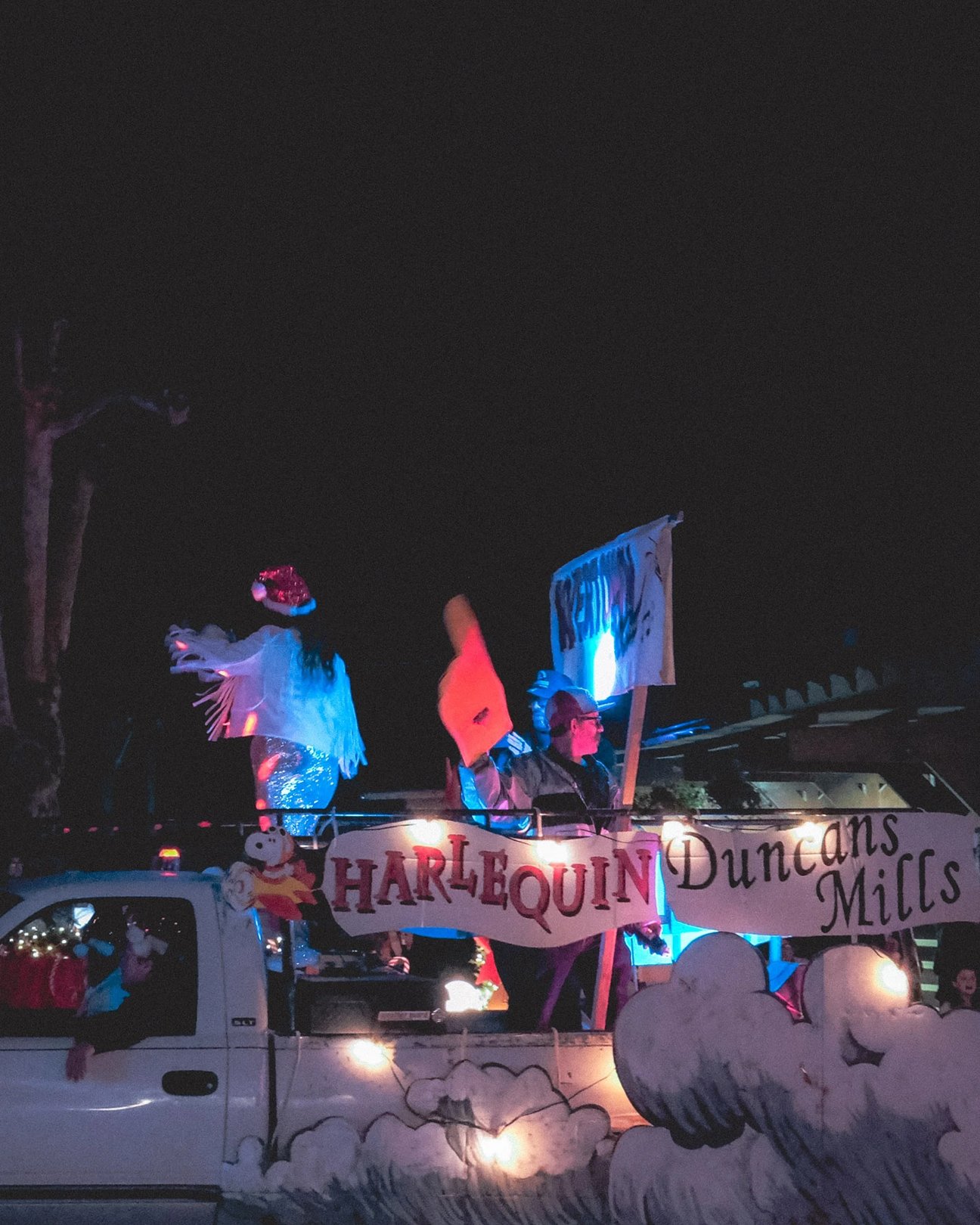 A decorated truck featuring performers holding a sign that reads "Harlequin Duncans Mills" in a festive nighttime parade.