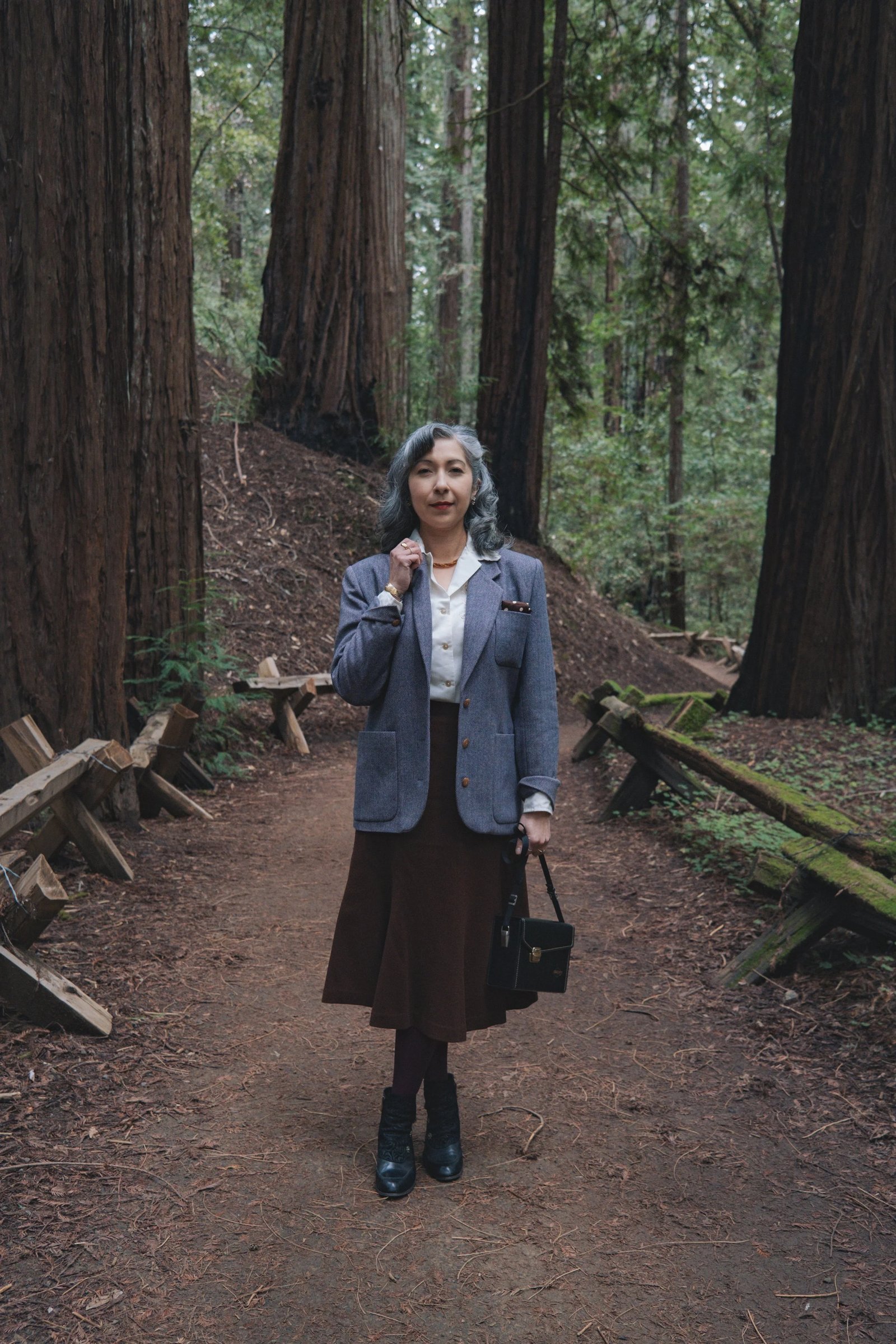 A woman in a gray blazer and brown skirt stands on a forest path surrounded by tall redwood trees.