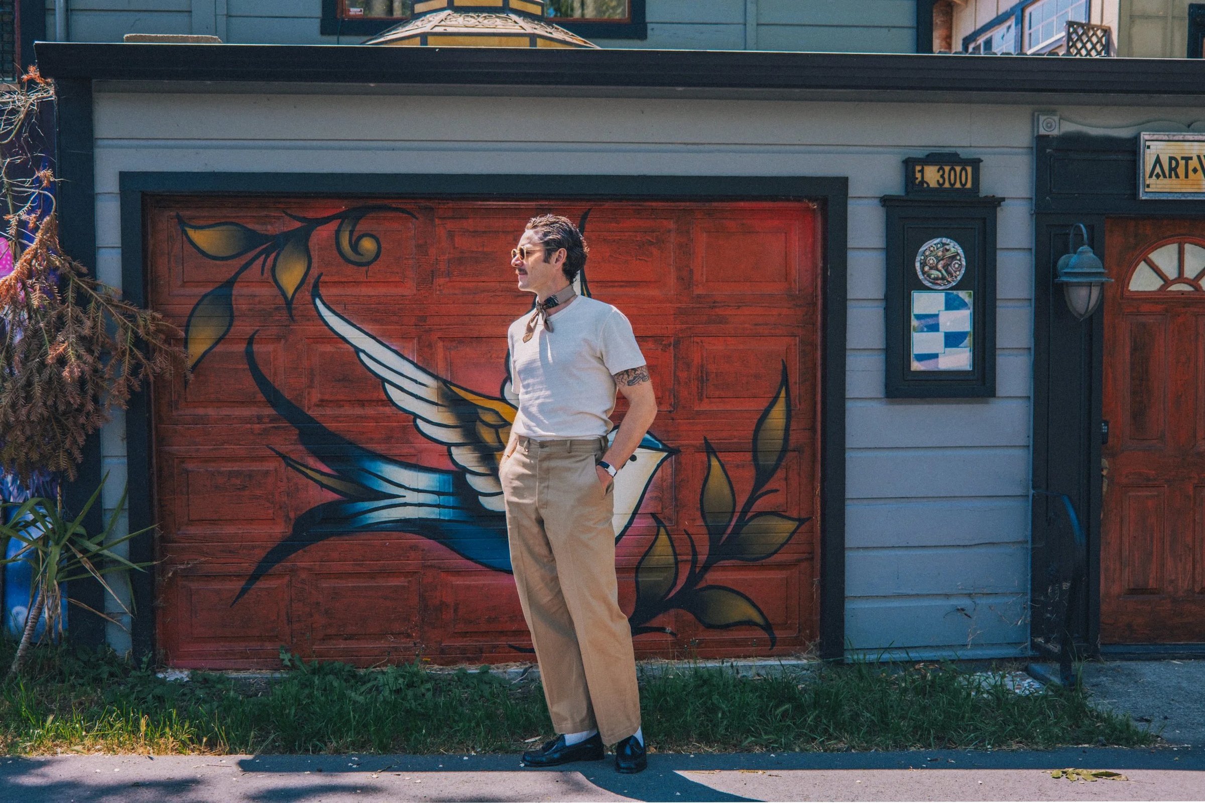 A person stands in front of a colorful mural, wearing a white shirt, beige trousers, and sunglasses, with a vintage hairstyle and a bow tie.