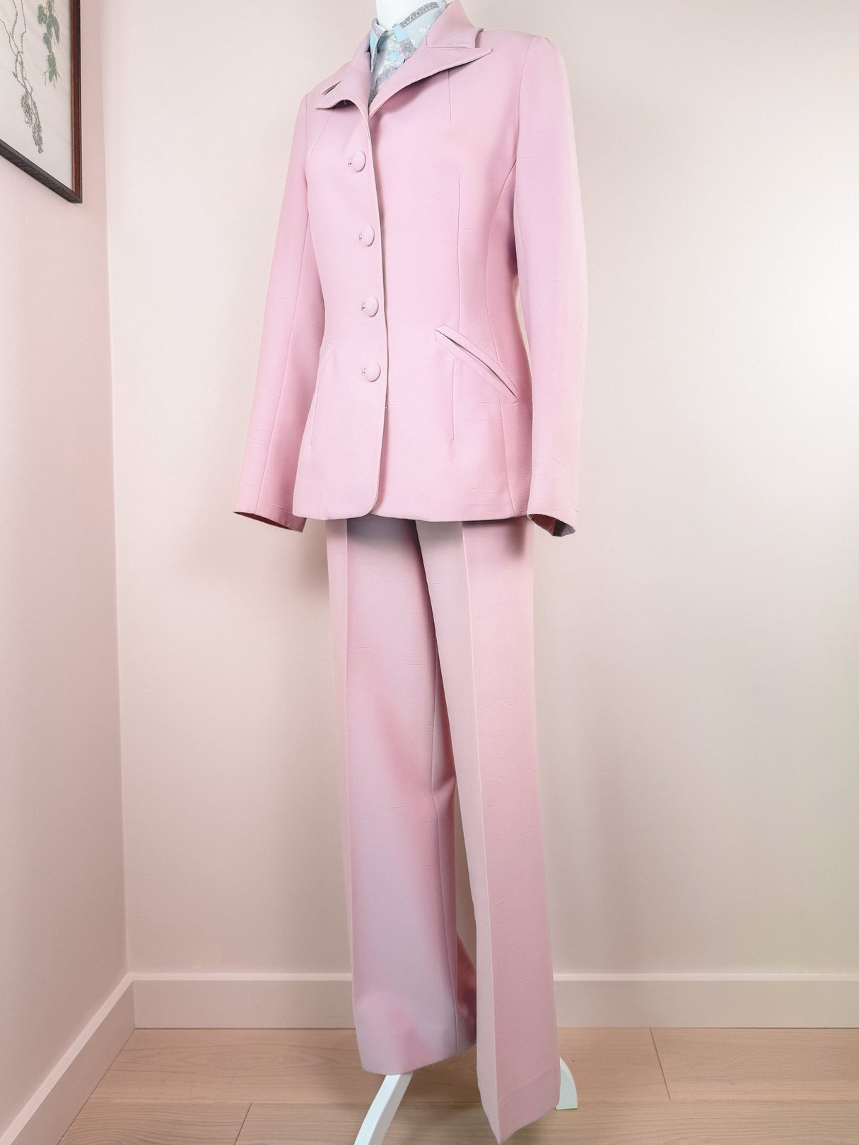 Blush pink three-piece women's suit from the 1970s, featuring a fitted jacket with buttons and matching trousers, displayed on a stand against a neutral background.