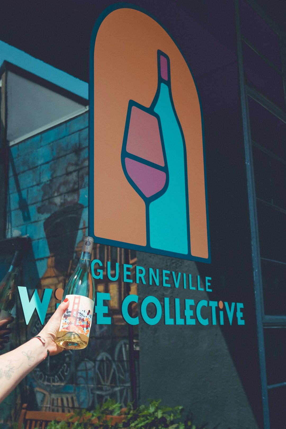 A person holding a wine bottle in front of the Guerneville Wine Collective mural featuring a wine bottle and glass.