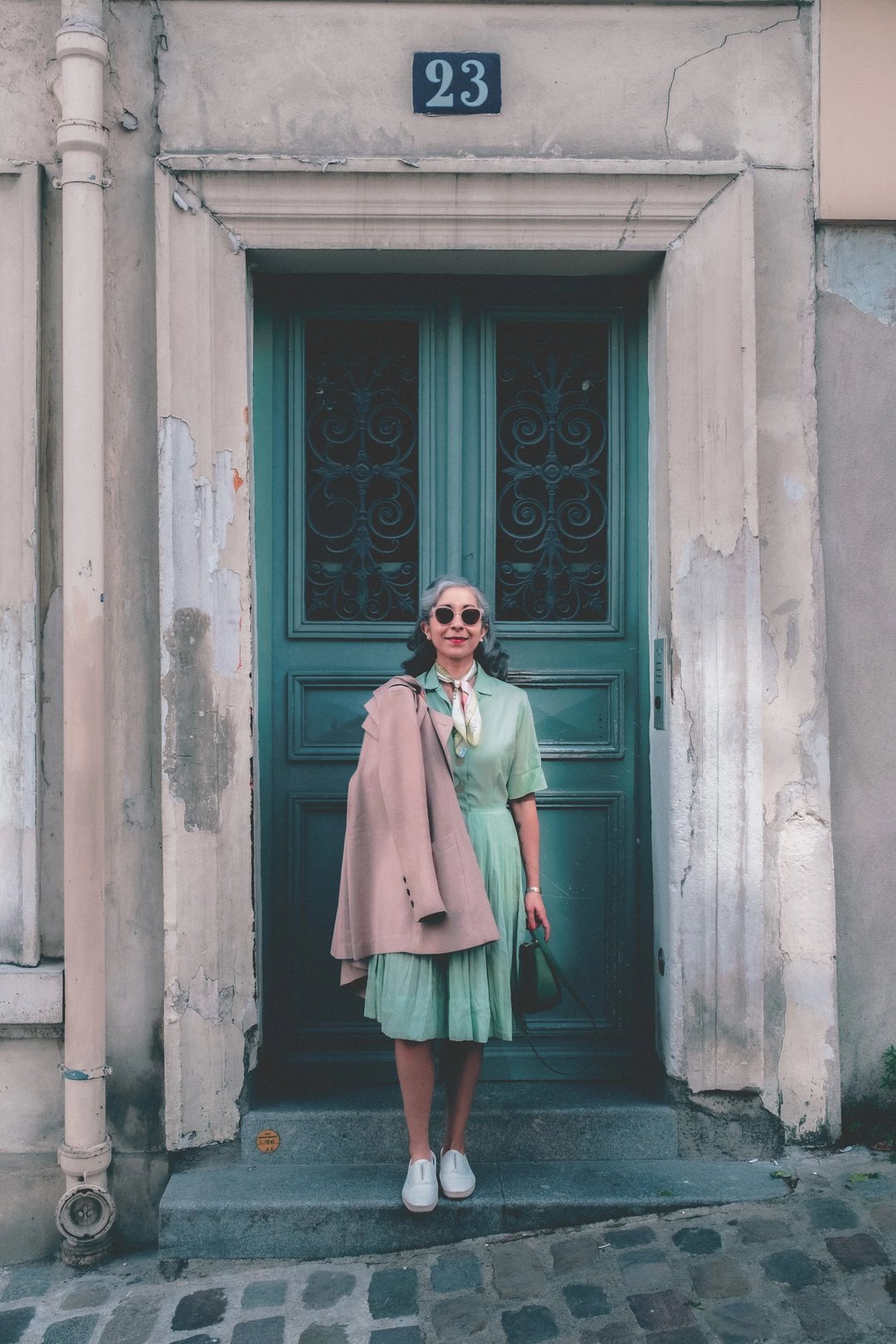 A woman in a mint green dress and sunglasses stands in front of an ornate green door, holding a tan jacket and a small handbag.