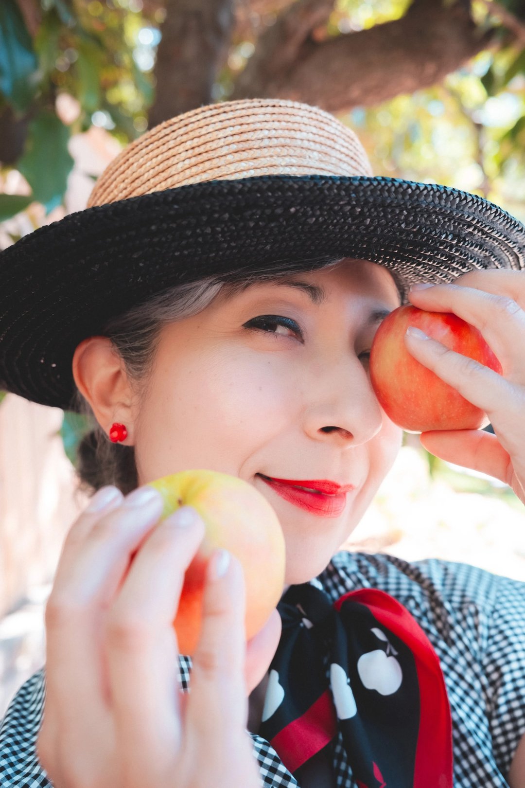 A woman wearing a straw hat and a vintage-style outfit holds two apples while smiling playfully.
