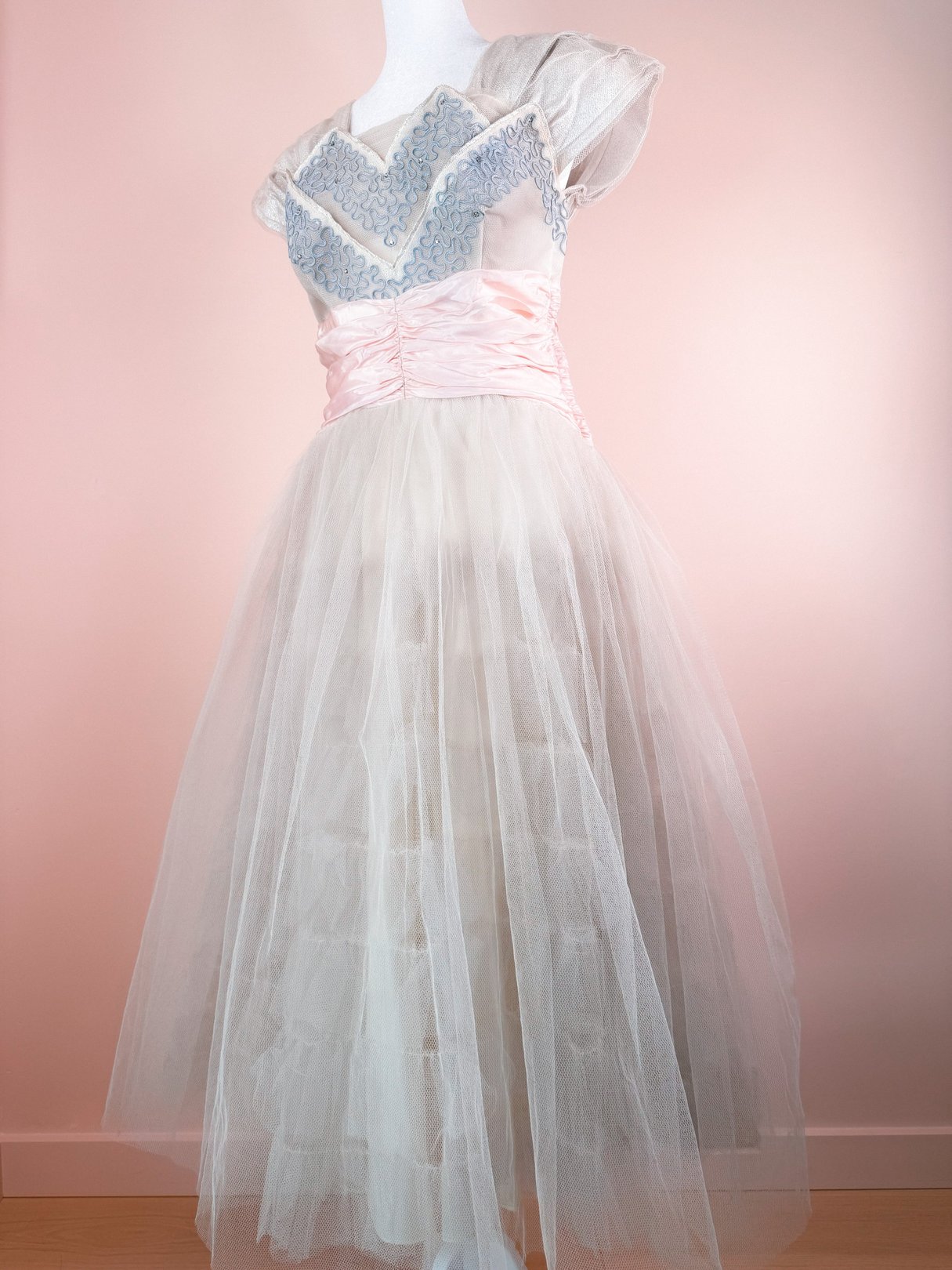 The Twilight Waltz Dress is a 1950s women's gown featuring a silver tulle skirt, pink satin waistband, and intricate blue embellishments on the bodice.