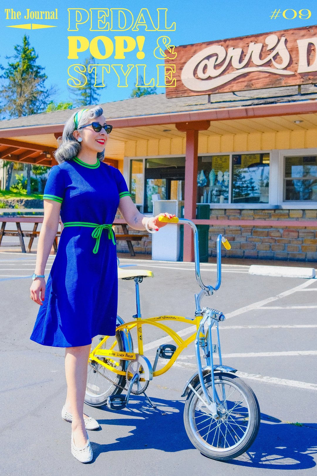 A woman in a blue dress with green accents walks beside a yellow bicycle in front of a retro diner.