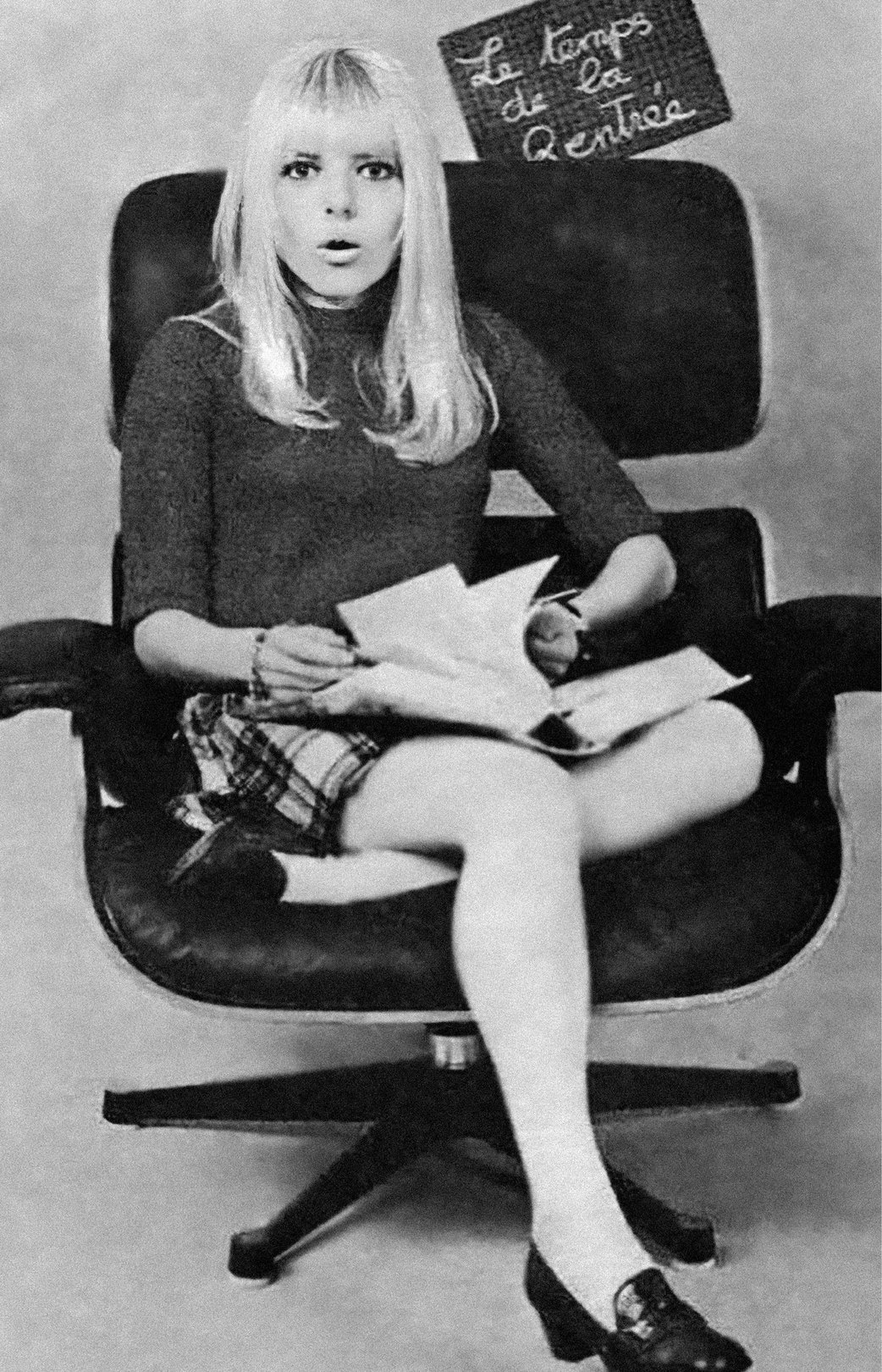 A woman with long blonde hair sits in a black leather chair, wearing a short plaid skirt and reading papers with a surprised expression.