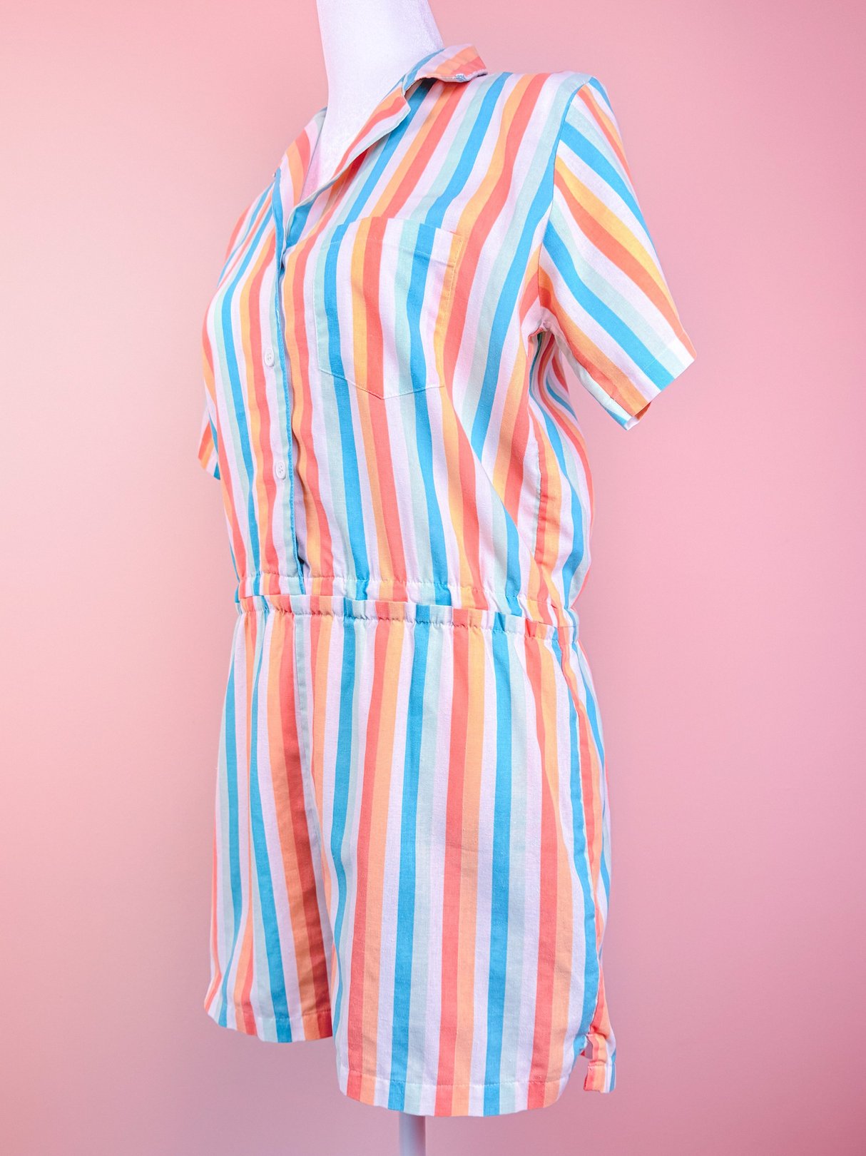 A women's 1980s striped romper in blue, orange, and pink, featuring a front pocket and short sleeves, displayed on a mannequin against a pink background.