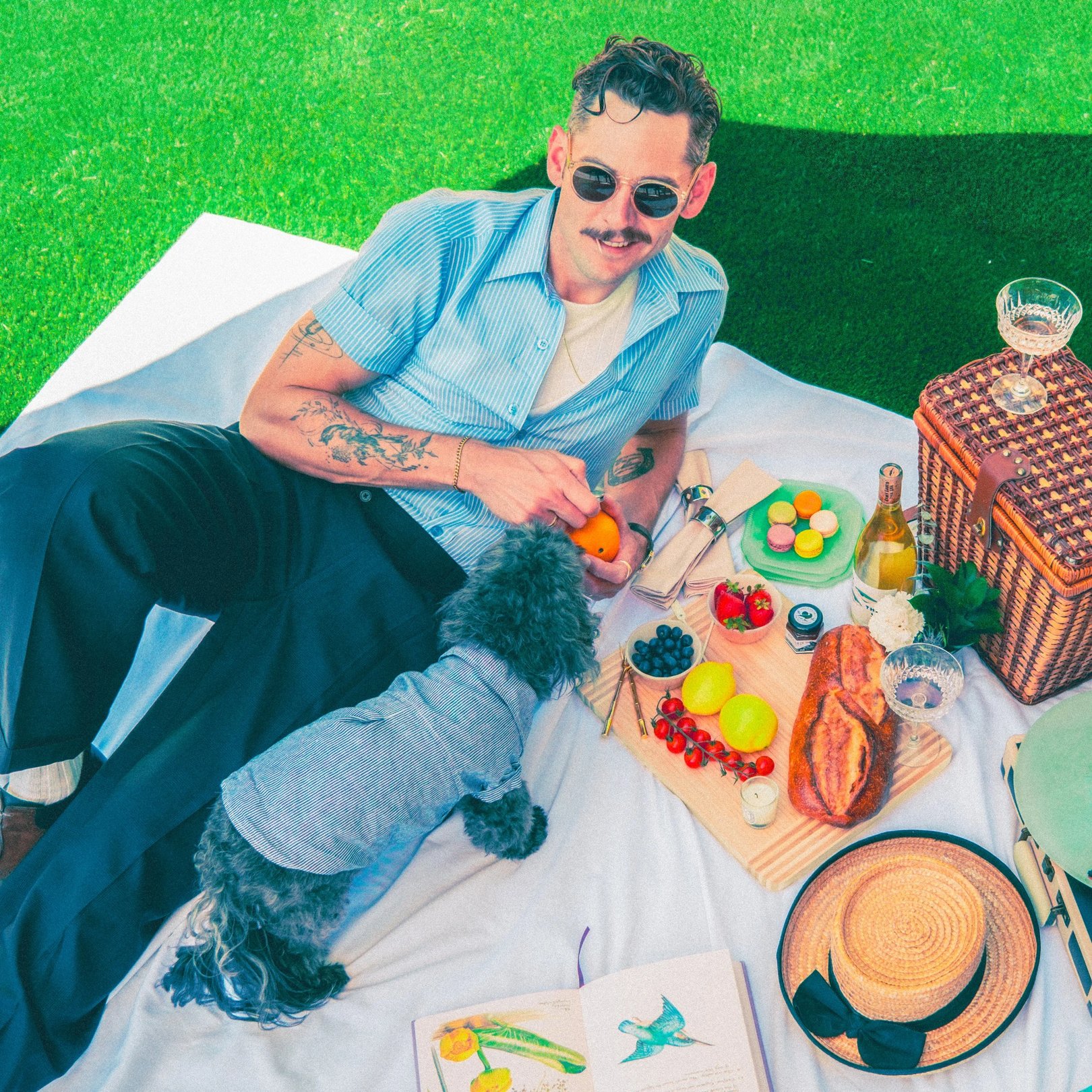 A man is seated on a picnic blanket with a variety of food items and a dog beside him.