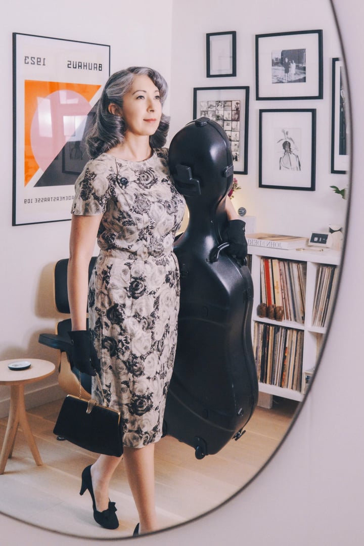 A woman in a floral dress and gloves carries a black cello case while walking through a stylishly decorated room with framed artwork.