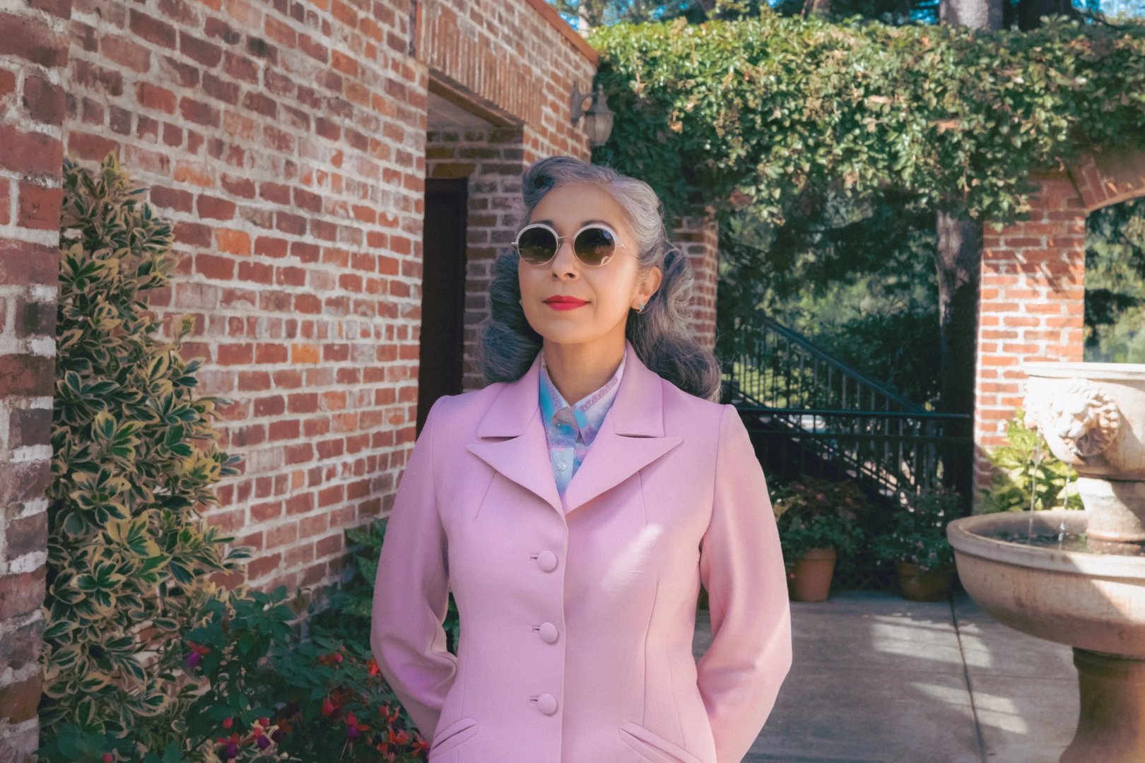 A woman in a pink suit and sunglasses stands outdoors near a brick wall and potted plants.