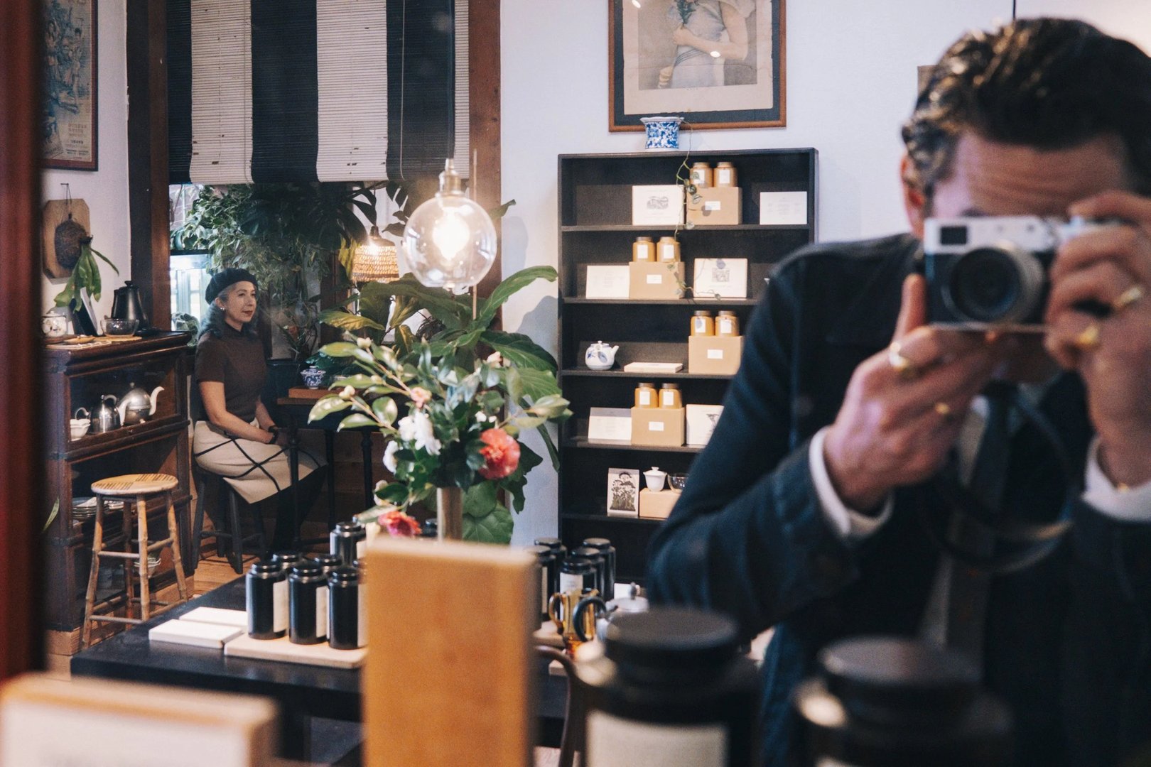 A man takes a photo with a camera in a shop, while a woman sits at a table in the background surrounded by plants and product displays.