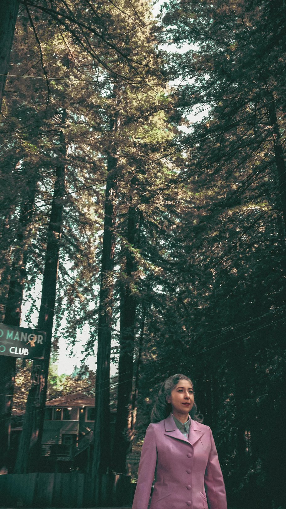 A woman in a lavender suit stands among tall redwood trees near a sign for a club.