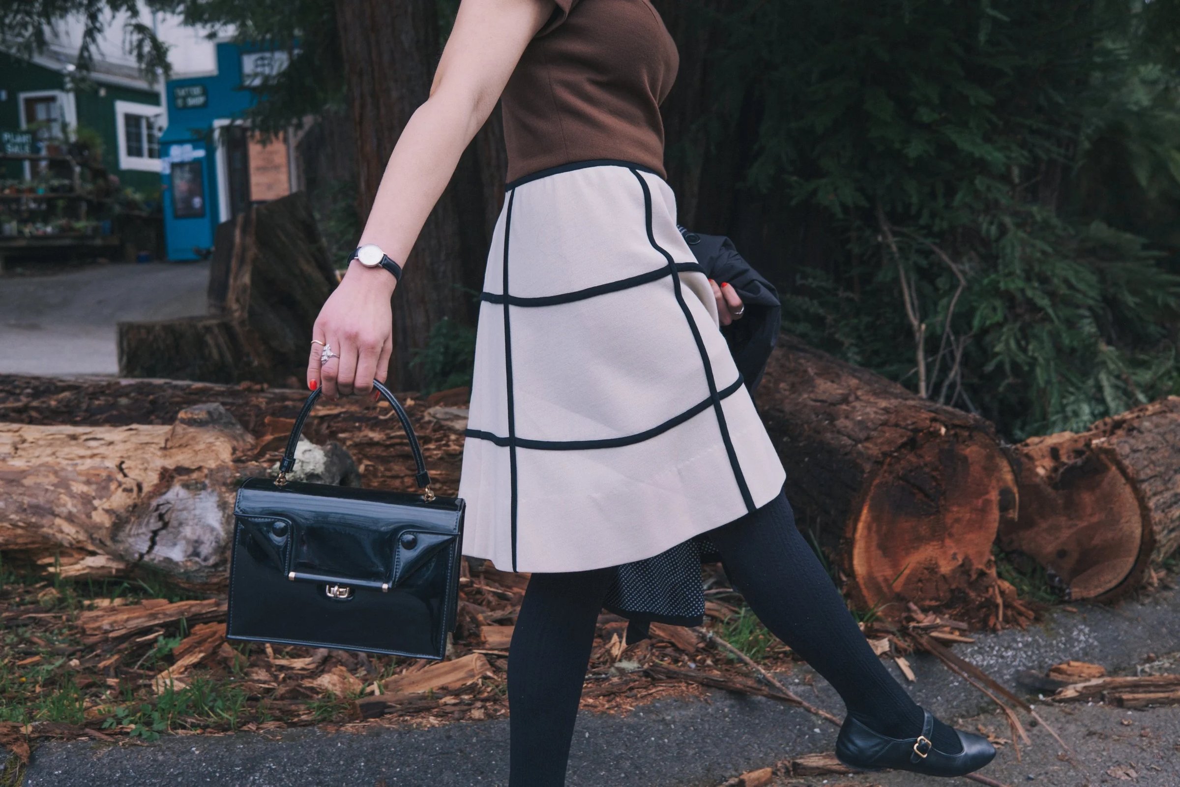 A woman walks in a brown top and a beige skirt with black stripes, carrying a black handbag.