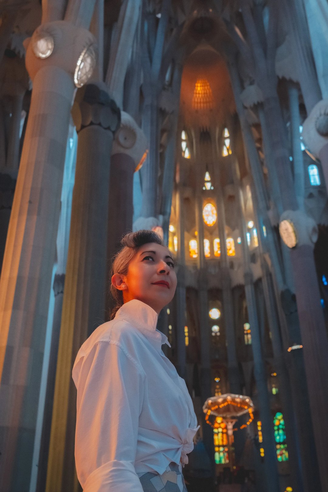 A woman stands inside the ornate interior of a gothic church with stained glass windows and towering columns.