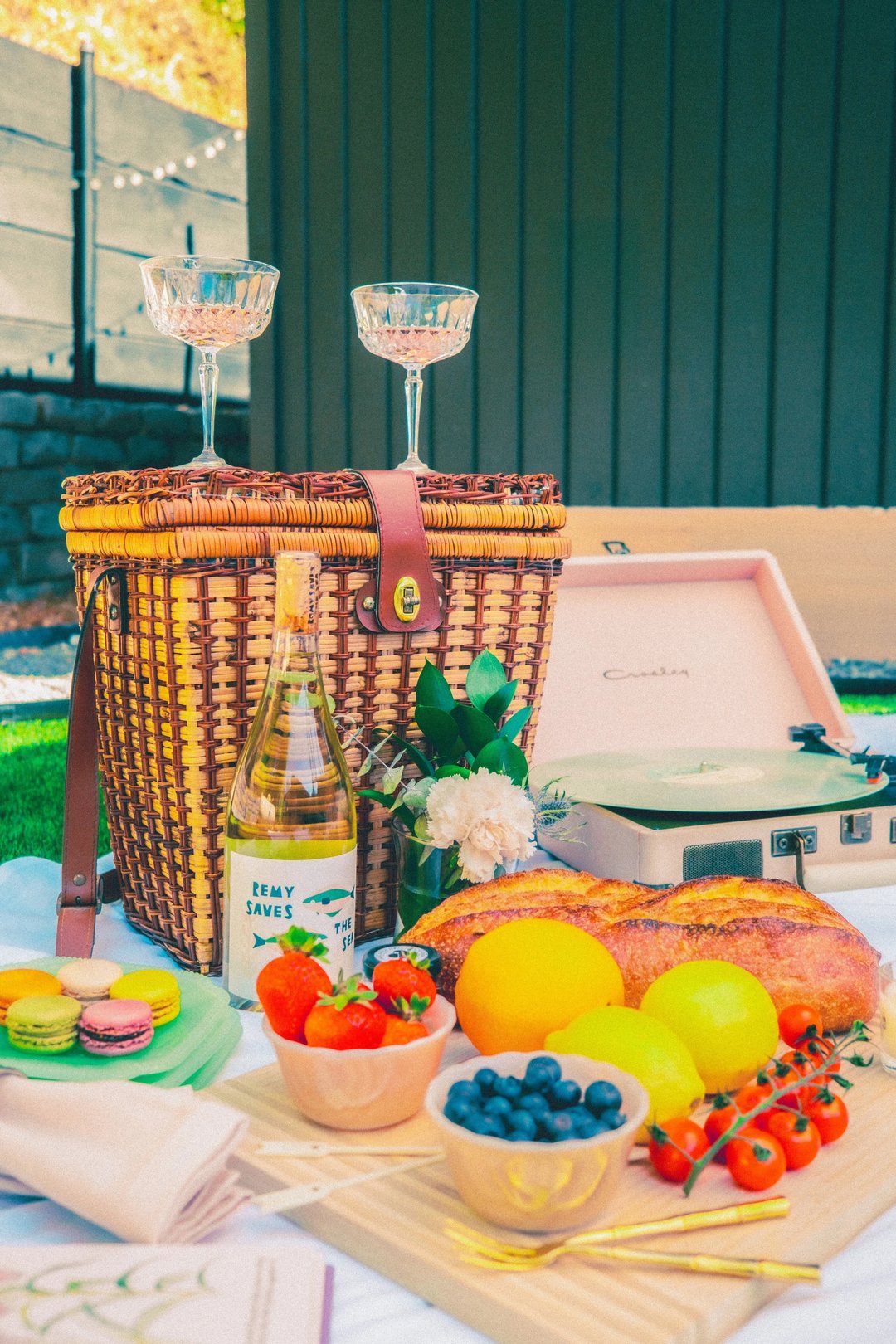 A picnic setup featuring a wicker basket, wine glasses, a bottle of wine, a record player, and a selection of fruits, macarons, and a baguette on a white blanket.