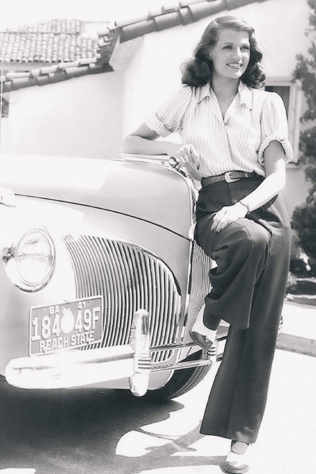 A woman wearing a striped blouse and high-waisted trousers poses stylishly next to a vintage car.