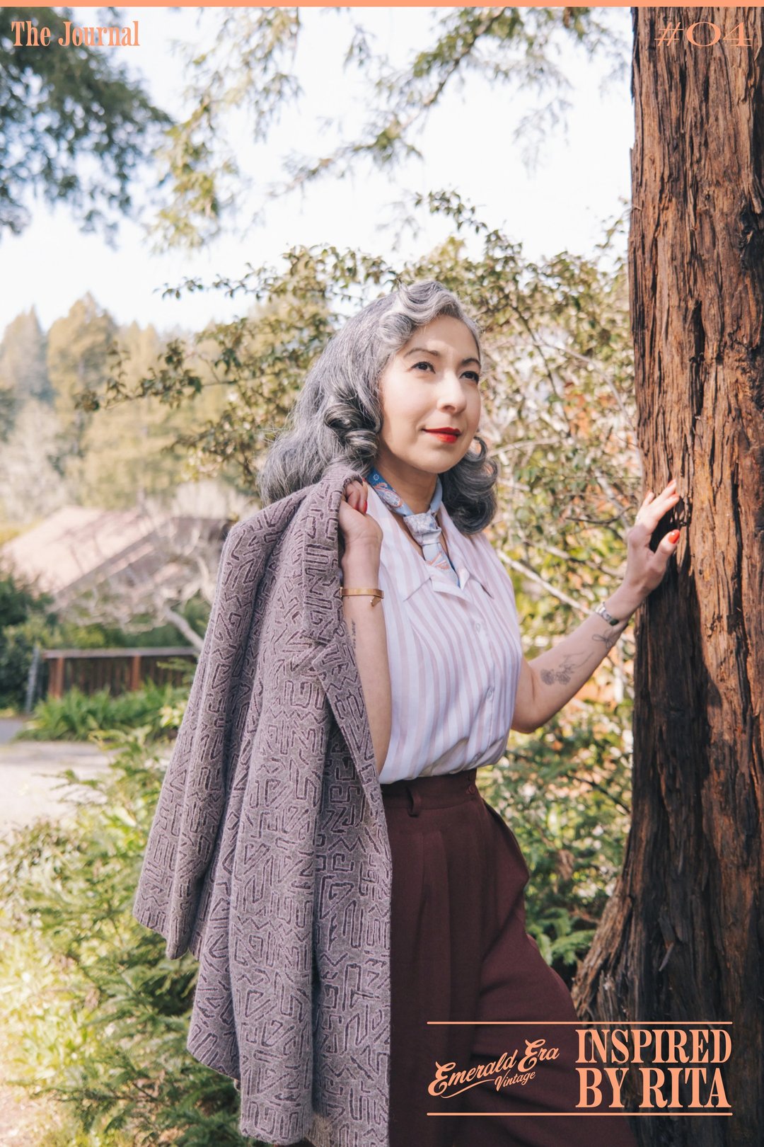 A woman with gray hair stands beside a tree, wearing a light blouse and a long gray cardigan with burgundy trousers.