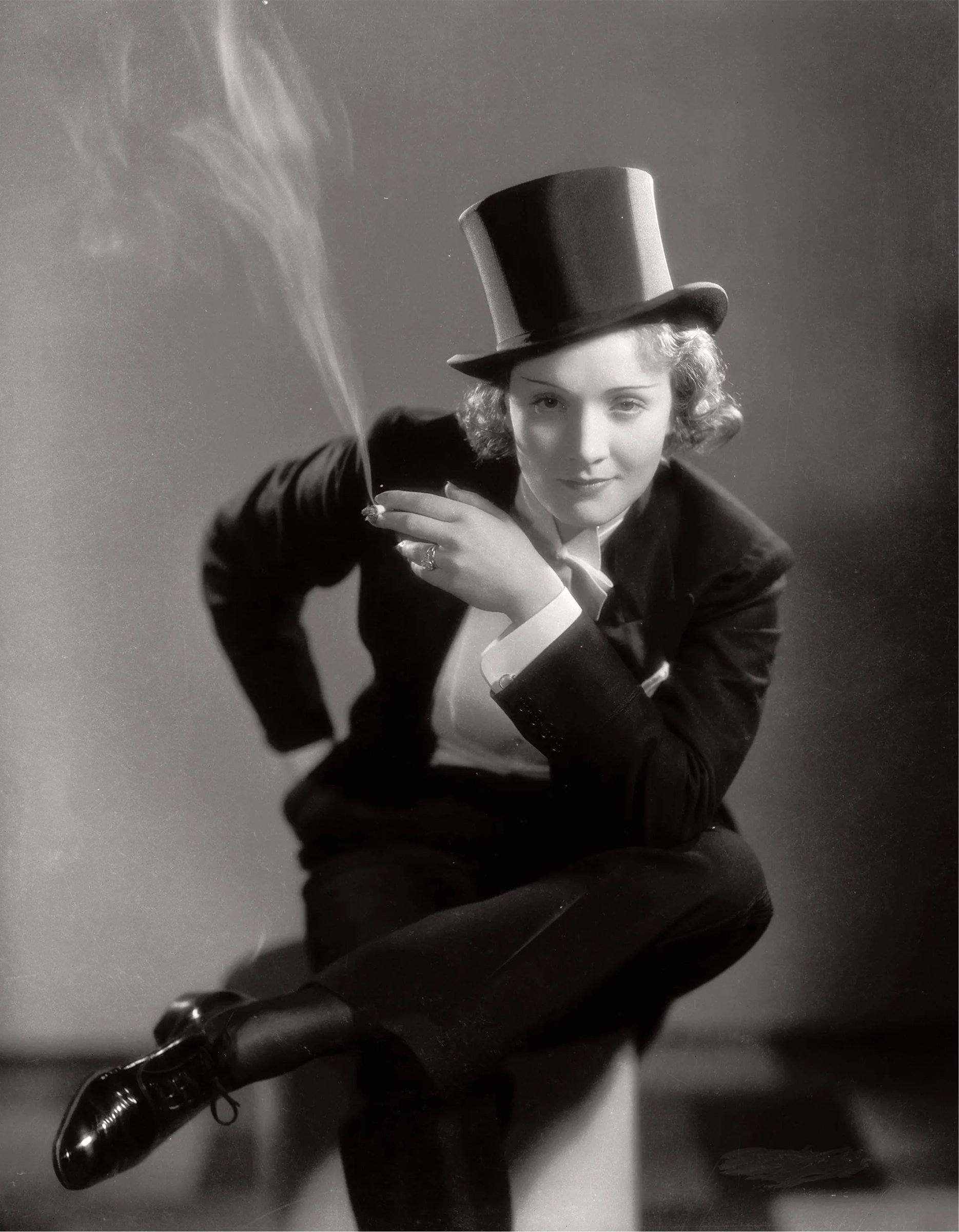 A woman in a formal suit and top hat sits casually, holding a cigarette with smoke curling upward.