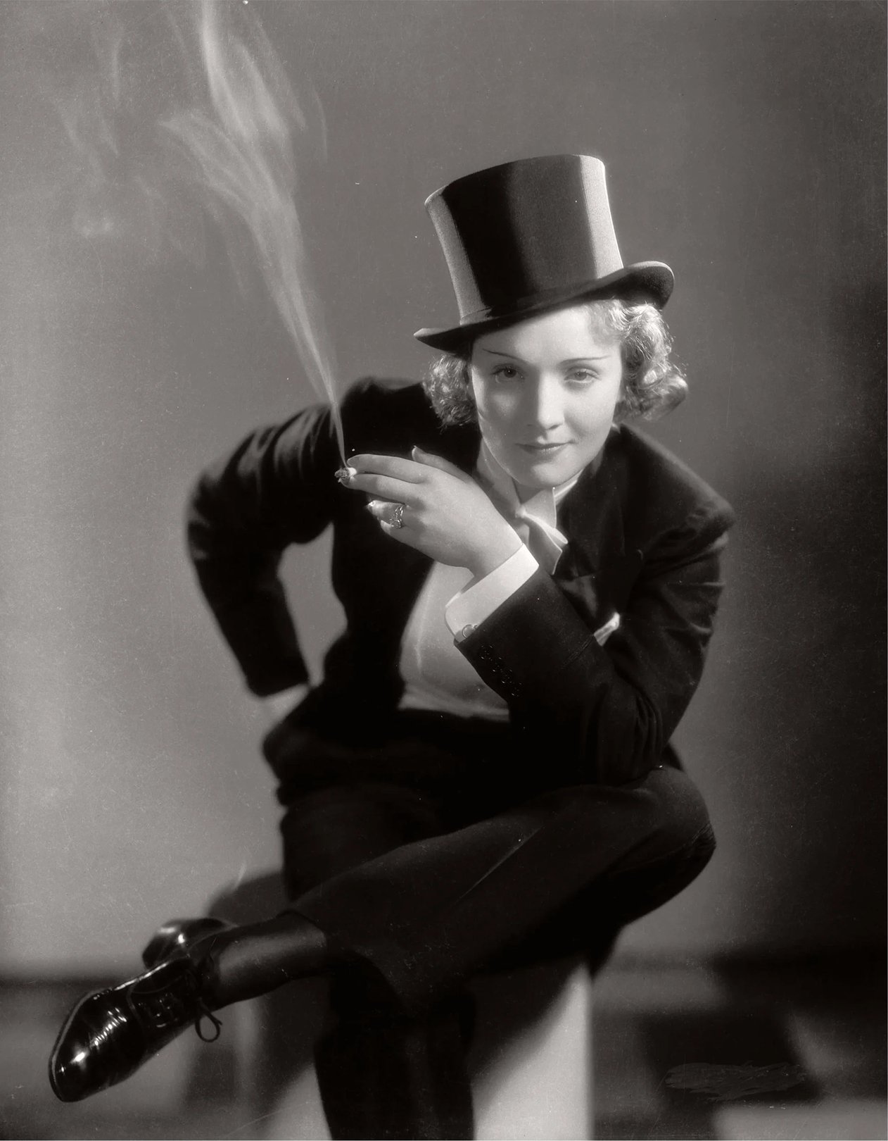 A woman in a formal suit and top hat sits casually, holding a cigarette with smoke curling upward.
