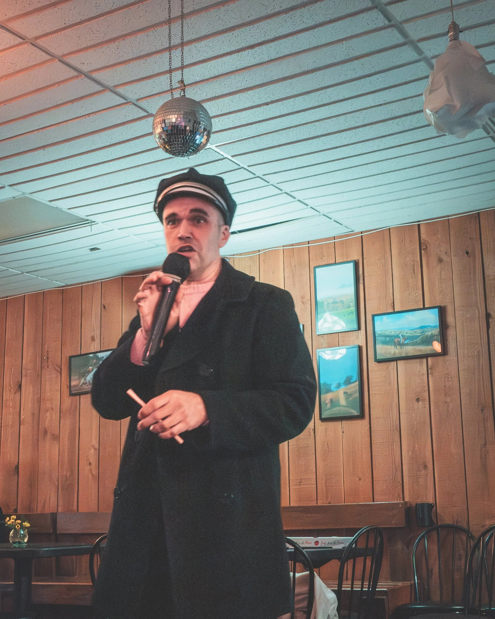 A performer in a captain's hat holding a microphone, standing in front of wooden walls adorned with framed paintings.