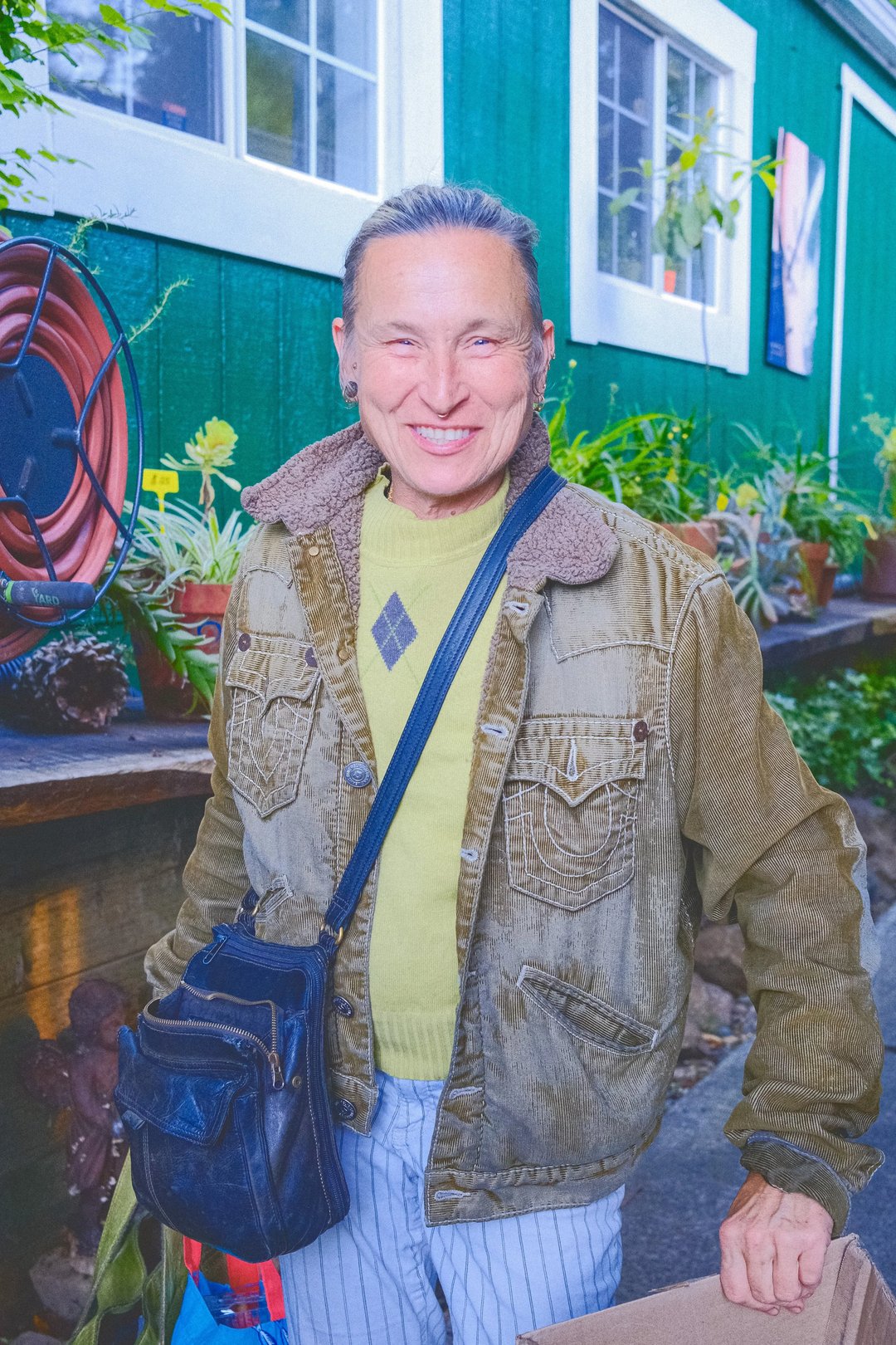 A smiling individual wearing a green sweater and a brown corduroy jacket stands outdoors in front of a green building adorned with plants.