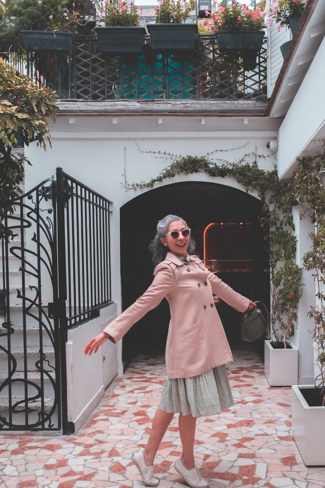 A woman in a pink coat and sunglasses joyfully poses in a charming courtyard with floral decorations.
