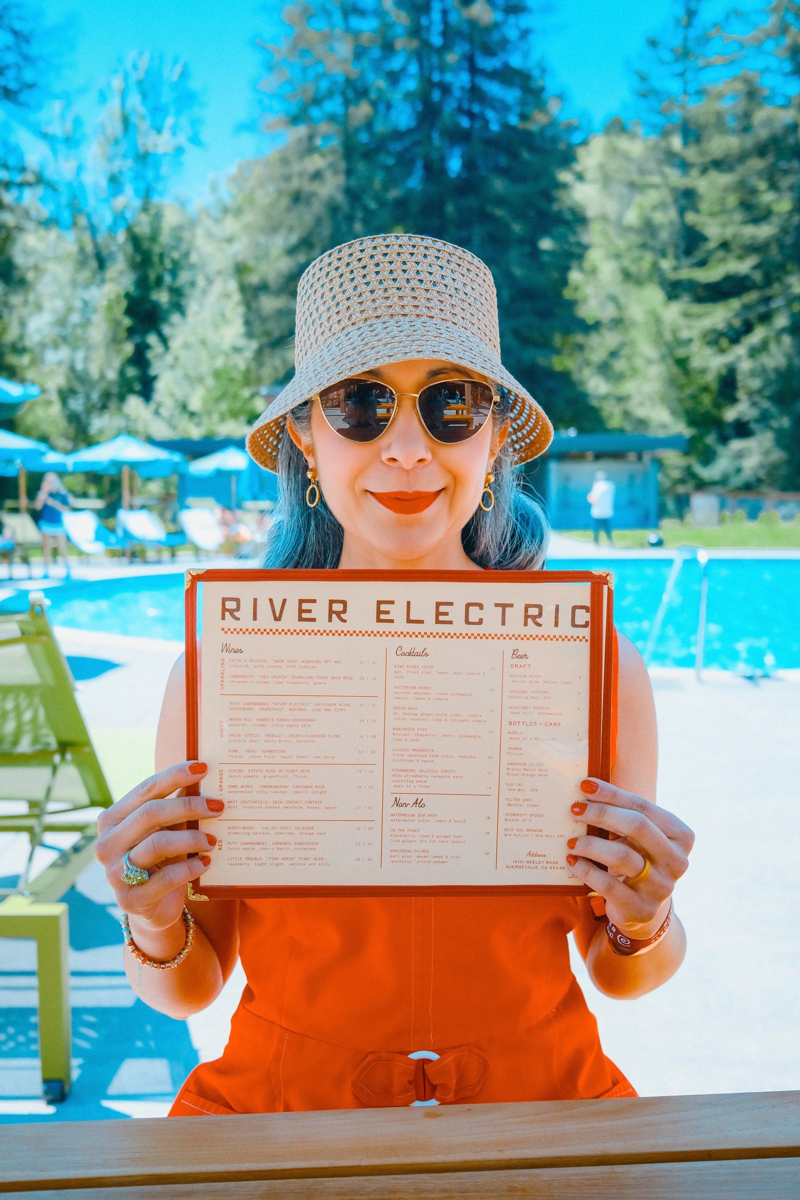 A woman in a straw hat and sunglasses holds a menu titled "River Electric" by a poolside setting.
