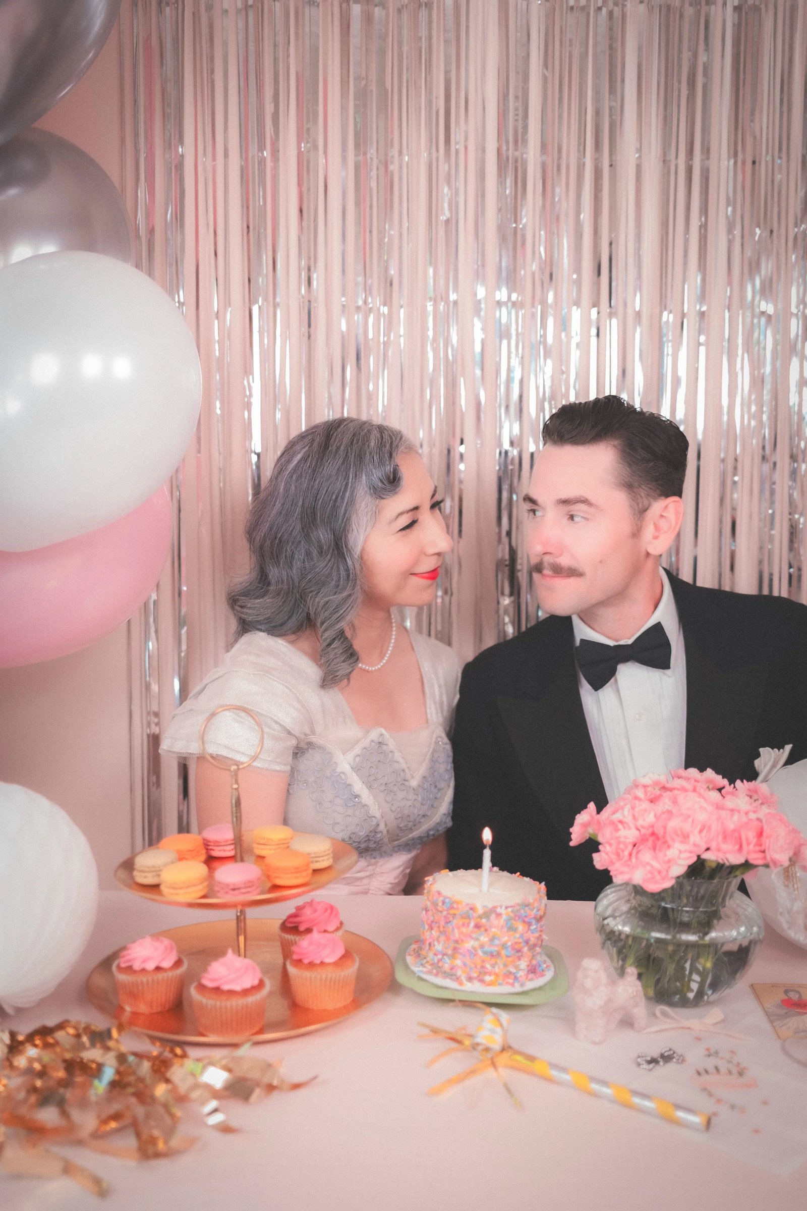 A couple dressed in vintage attire sits at a table decorated with a colorful cake, cupcakes, flowers, and balloons, set against a festive backdrop.
