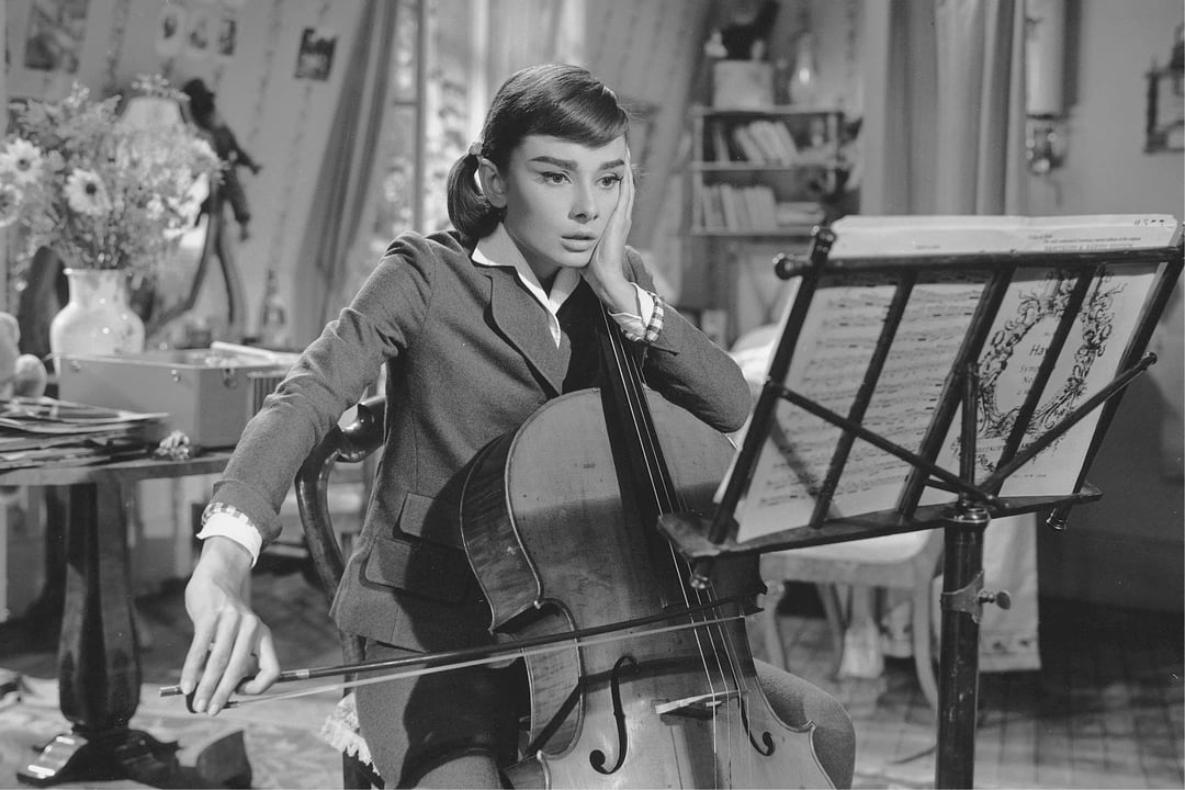 A young woman in a vintage outfit plays the cello while looking thoughtfully at music sheets in a cozy indoor setting.