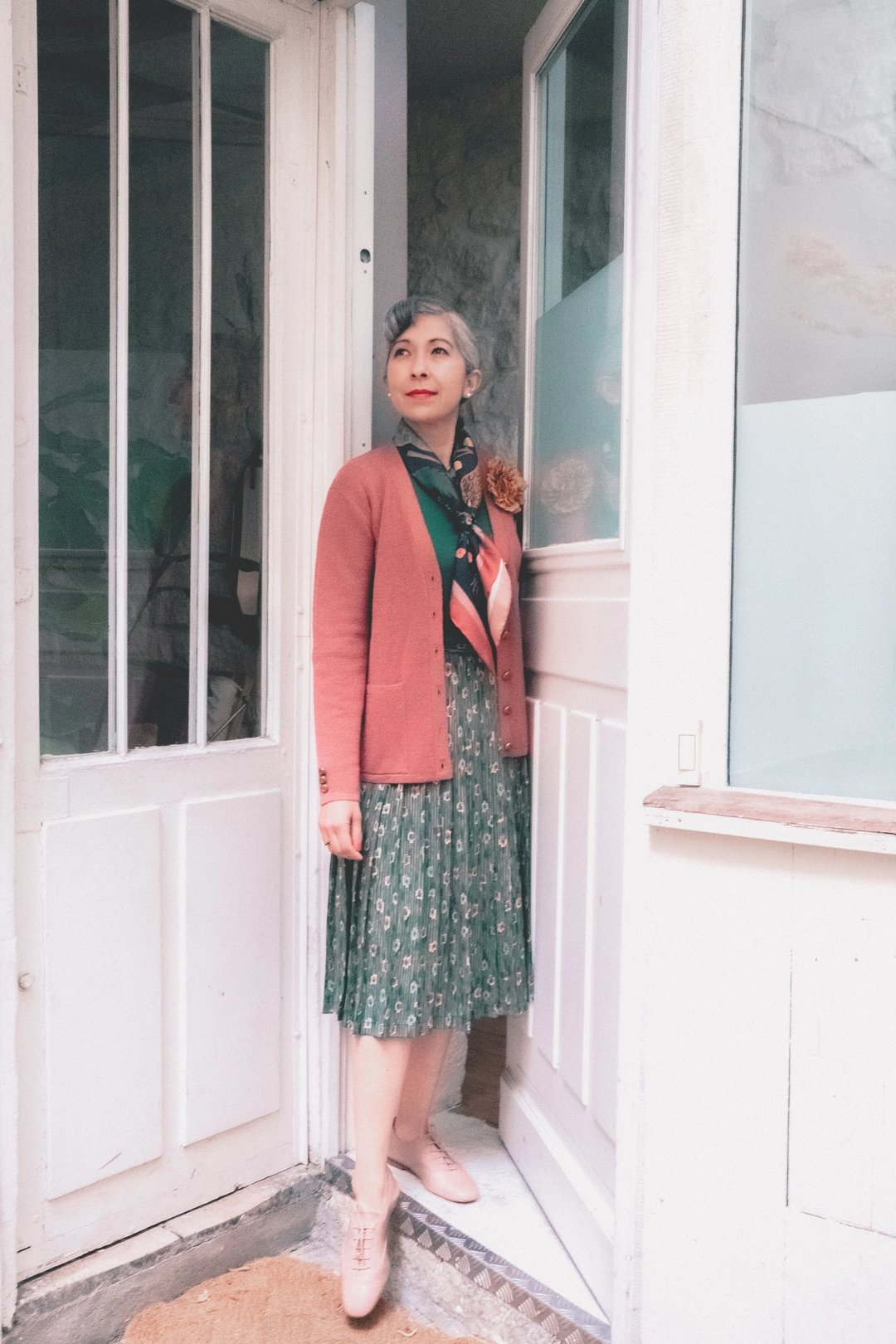 A stylish woman stands at the entrance of a doorway, wearing a green floral dress, a pink cardigan, and pastel shoes, with a flower accessory in her hair.
